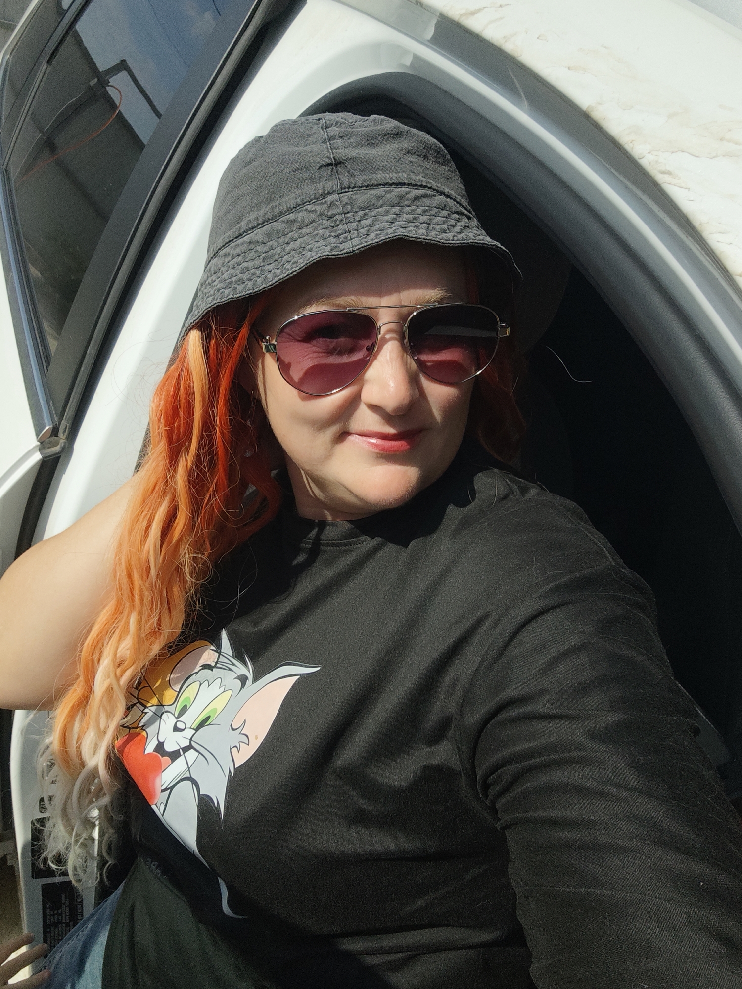 The image shows a woman with bright orange hair and sunglasses, wearing a bucket hat and a black t-shirt with a cartoon cat graphic. She is seated inside a white vehicle, with the door open to reveal a sunlit environment. The background suggests an outdoor setting, possibly in a rural or semi-urban area, given the appearance of a building and sky. There is no direct indication of the specific location within the city of Mokino, Russia, as no landmarks or specific geographical features are visible. The woman appears to be posing for a selfie, with her arm resting on the car's interior.