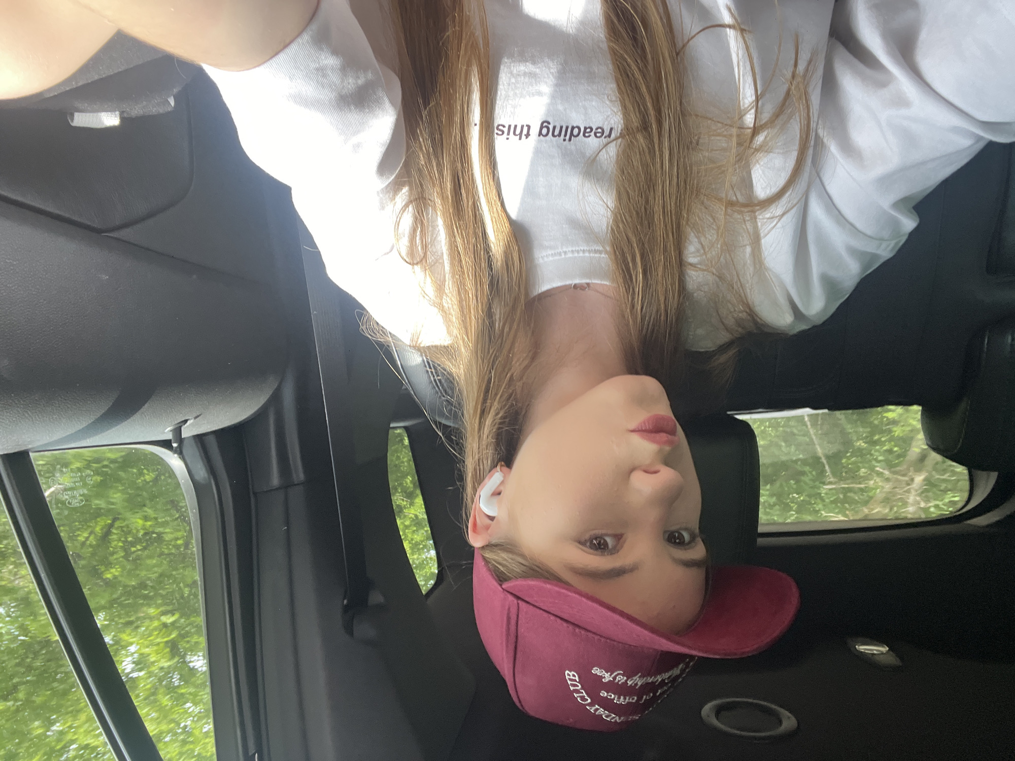 A young woman with long blonde hair is taking a selfie inside a car. She is wearing a white t-shirt with black text that reads "reading this." She has on a maroon baseball cap with white embroidered text that appears to say "SUNDAY CLUB." She is also wearing white wireless earbuds. Her expression is neutral, and she is looking at the camera. The background shows trees and foliage, indicating it is daytime and likely pleasant weather. The interior of the car is visible, with dark upholstery and a car door. The image is oriented upside down.