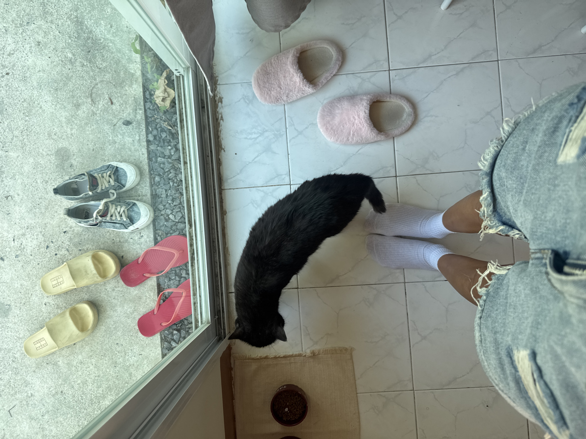 A high-angle view from inside a building in Choeng Thale, Thailand, shows an interior tiled floor transitioning to an exterior concrete surface through a doorway. The lower body of a person, wearing distressed blue denim shorts and white ankle socks, is visible on the light-colored tiled floor. A black cat stands nearby, its head lowered towards two brown bowls on a beige mat, one containing dry food. A pair of pink, fuzzy slippers rests on the tiles to the right. Beyond the white door frame, on the concrete ground, are three pairs of shoes: cream-colored slide sandals, red flip-flops on a gravel strip directly outside the door, and faded blue canvas sneakers. The gravel strip includes minor debris, and sparse green vegetation is present on the exterior concrete.