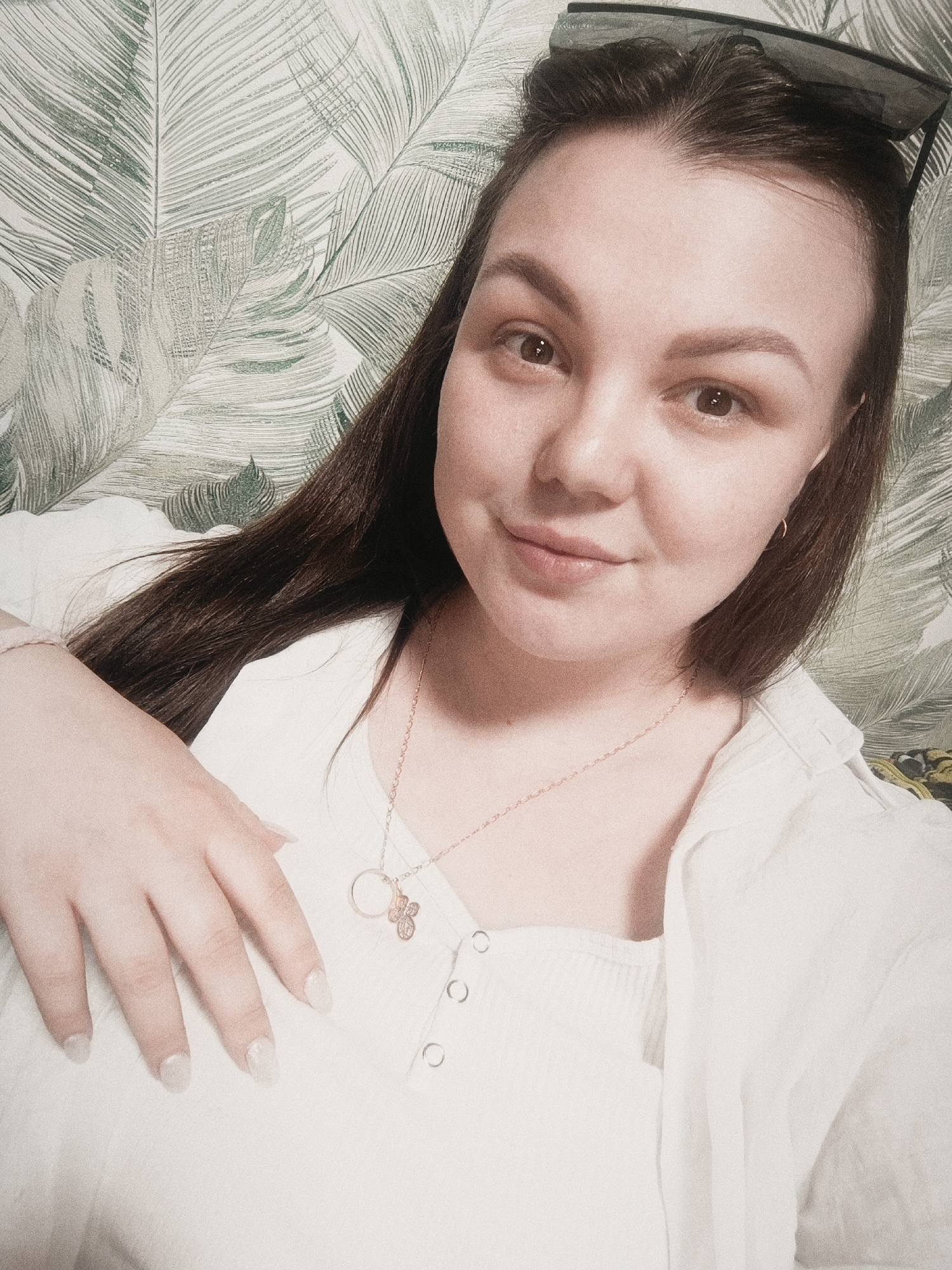 A woman is posing for a selfie. She has brown hair, fair skin, and is wearing a white shirt. She is also wearing a gold necklace with a butterfly pendant. Sunglasses are perched on top of her head. The background features a wallpaper with a leaf pattern. The location is likely indoors, but the exact location in the city of Прикамский, Russia is not identifiable from the image.