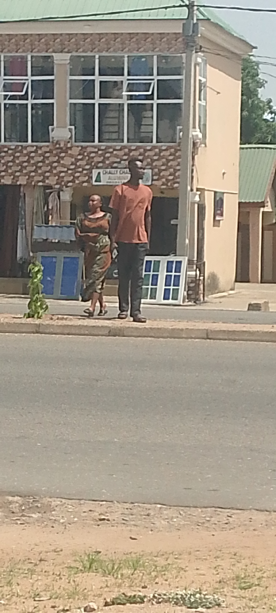 A full shot shows a man and a woman standing on a sidewalk in Jalingo, Nigeria, in front of a building with multiple windows. The building has a light tan facade with a patterned brick base. Above the brick base, there are multiple rows of windows with white frames. Some of the windows are open, revealing the interior of the building, which appears to be a storefront. A sign with green lettering that reads "CHALLY CHALUMINUM" is visible above the entrance. The man is wearing an orange t-shirt and dark pants, and the woman is wearing a patterned dress. Both are standing still and facing to the right, looking at something outside the frame. In front of them on the sidewalk are two large window frames, one with blue panes and another with blue and green panes. A small, leafy green plant grows in a pot to the left of the man and woman. The sidewalk is made of concrete, and behind it is a paved road. The background shows more buildings with green roofs and some trees. The sky is bright and clear. The image appears to have been taken during the daytime.