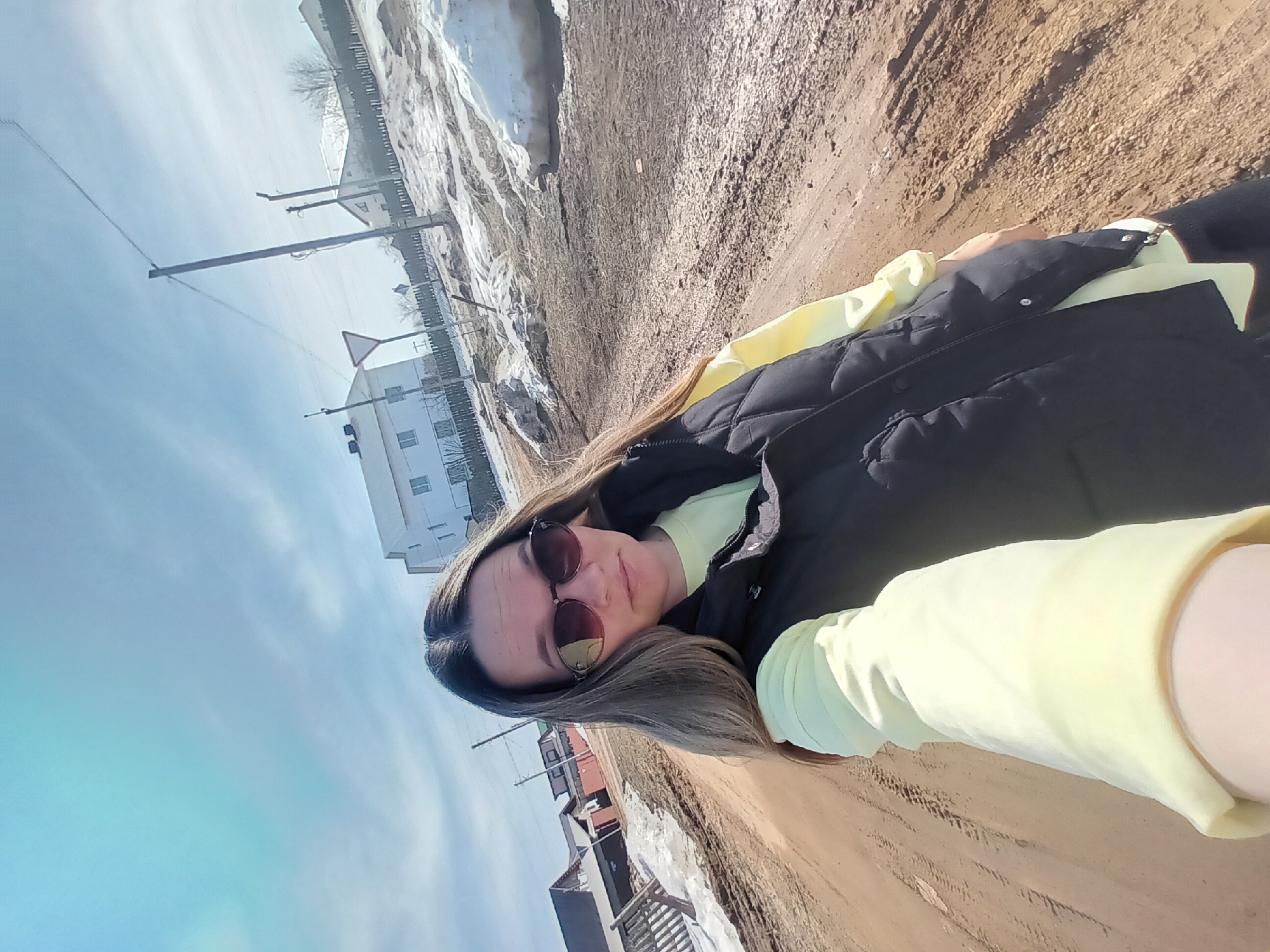 A woman is seen outdoors on a dirt road in what appears to be the city of Ольховка, Russia. She wears sunglasses, a black vest over a pale yellow shirt, and her hair is long and light brown. The road is bordered by snow and buildings, with a bright sky above. A utility pole is visible near a building.