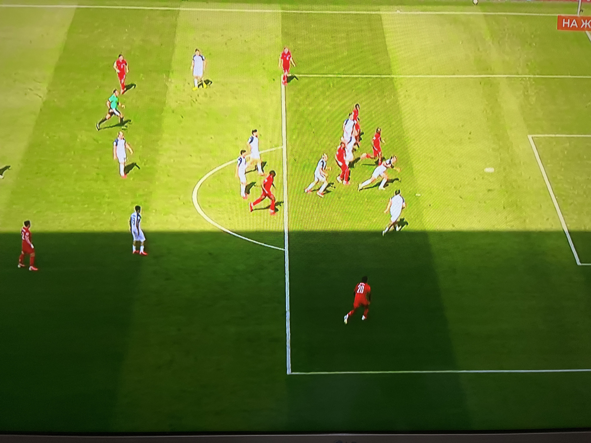 This is an image of a soccer match being played on a grass field. Several players are visible on the field, wearing red and white uniforms. The players are in various positions, some running, some standing, and some in the process of kicking or receiving a ball. The field is marked with white lines, indicating the boundaries of the playing area. There are shadows cast on the field, suggesting it is daytime and the sun is shining. A portion of a goalpost is visible in the lower right corner of the image. There is also a sign with Russian text "НА Ж" visible in the upper right corner of the image. The scene is an outdoor sporting event.
