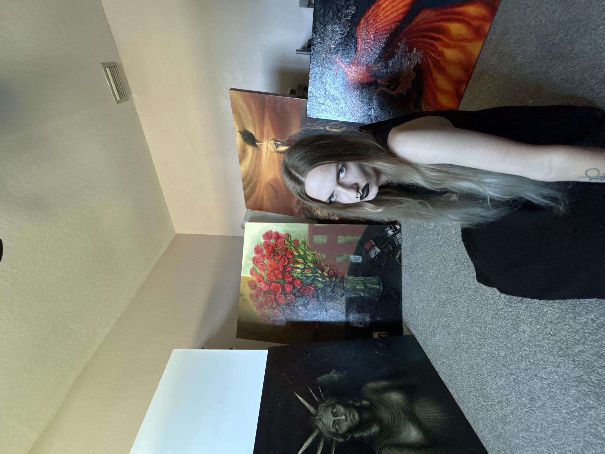 A young woman with long, light blonde hair, dark lipstick, and a septum piercing is prominently featured in a room filled with several large paintings. She wears a black top, and a tattoo is visible on her left forearm. Her expression is serious and direct, with her head tilted as she looks towards the viewer.
The room has light-colored walls, one of which features a rectangular air vent. The floor is covered with a grey, textured carpet.
Several diverse paintings are displayed around the woman:
1. Behind her head, a vibrant painting depicts a fiery orange and red creature, resembling a phoenix, against a dark, textured background.
2. To her left, another painting with warm, earthy tones suggests a stylized figure or dramatic landscape.
3. Below the fiery painting, a still life artwork showcases a large bouquet of red roses in a vase on a dark surface. Within this painting, bottle-like objects with the visible text "MEXICO COKE" are present in the background.
4. At the bottom of the frame, a dark, monochromatic painting portrays a somber figure resembling the Statue of Liberty or a similar symbolic entity, complete with a spiky crown, set against a starry sky.
The woman appears to be posing among her artwork or a collection of paintings. The lighting suggests it is daytime. The scene is set indoors, in what appears to be a home or studio in Cape Coral, United States of America.