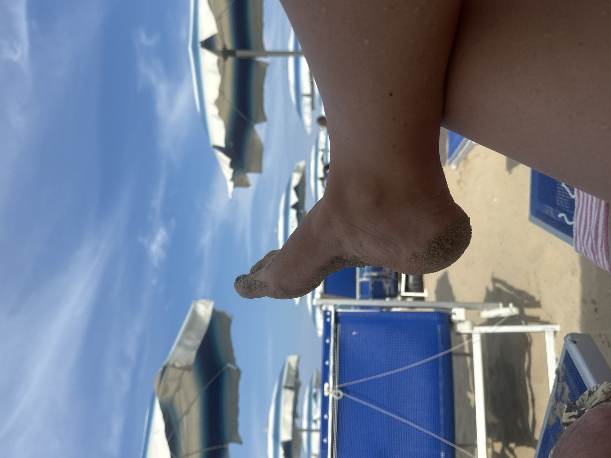 A person's sand-covered foot and lower leg are the main subject of this image, taken from a low-angle perspective. The foot is dirty with sand, particularly around the heel and toes. The skin appears sun-kissed. In the background, several beach umbrellas with blue and white stripes are visible, some tilted. The sky is bright blue with scattered white clouds, suggesting a sunny day. Below the umbrellas, blue beach chairs are lined up on a sandy beach. The image is oriented vertically. Given the location context of Pineto, Italy, and the presence of beach umbrellas and chairs, it is likely a scene on an Italian beach during a sunny day, possibly in the summer. The focus on the foot suggests a relaxed and perhaps post-beach activity moment. No text is visible in the image.