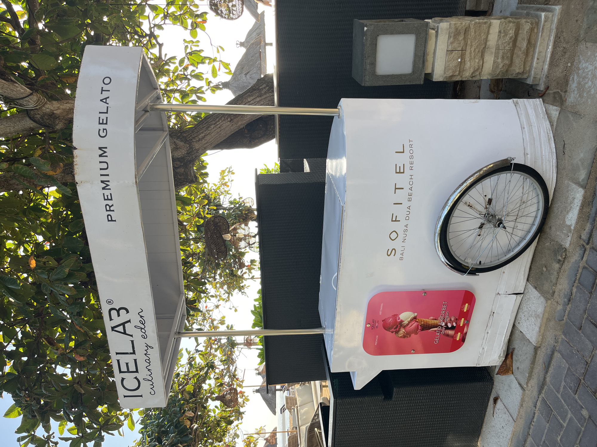 A white gelato cart with a metal frame is positioned on a paved walkway in front of a black woven wall. The cart has a white canopy with "ICELAB3 culinary eden" on the left and "PREMIUM GELATO" on the right. On the side of the cart, a pink rectangular sign displays an image of a three-scoop gelato cone and the text "GELATO & SORBET daily vegan." Below the sign, the text "SOFITEL BALI NUSA DUA BEACH RESORT" is visible. A bicycle wheel with a fender is attached to the cart. To the right of the cart, a square light fixture is mounted on a stone base. In the background, lush green trees with dappled sunlight are visible, along with a glimpse of a beach with lounge chairs and umbrellas. The ground consists of a mixture of light gray paving stones and dirt.