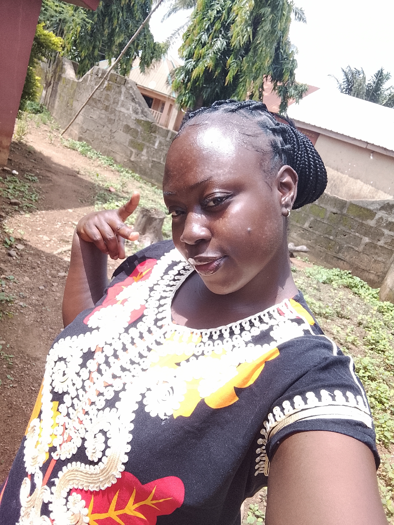 A young woman with braided hair is posing for a selfie outdoors in Akwanga, Nigeria. She is wearing a black t-shirt with a white, yellow, and red floral pattern. Her skin is dark and shiny, and she has on subtle makeup, including lip gloss. She is looking towards the camera with a slight smile and her left hand is raised, with her index finger pointing. Behind her, there is a dirt path, some low-lying green plants, and a stone wall. Further in the background, there are trees and buildings with white roofs. The lighting suggests it is daytime.