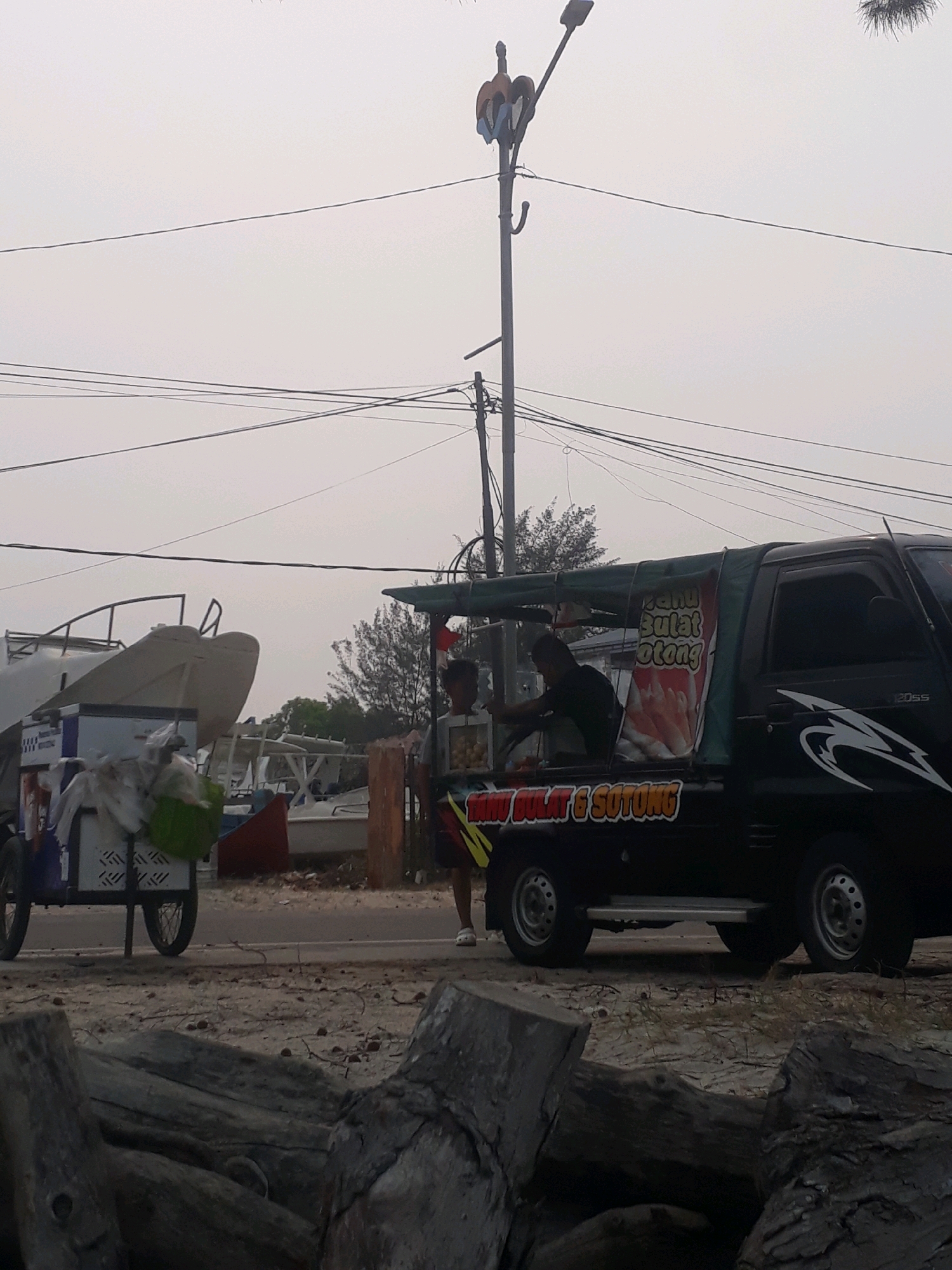 This image depicts a scene on a street in Pangkalpinang, Indonesia. In the foreground, large pieces of driftwood are scattered on a sandy surface. Beyond this, a paved road runs horizontally. On the left side of the road, a white and blue food cart is parked, piled with various items, including plastic bags and what appears to be produce. Behind the cart, several boats are visible, some appearing to be under construction or repair.
Further down the road, a black pickup truck is parked, adapted as a mobile food vendor. The truck has a green canopy extending over its bed, displaying signage that reads "TAU BULAT & SOTONG" in red and white lettering. A man in a black shirt is visible inside the truck's serving area, interacting with a customer. Another person, wearing shorts and white footwear, stands to the right of the truck.
In the background, a street light with decorative elements is mounted on a pole. Overhead, power lines crisscross the hazy sky. The environment appears to be a coastal or roadside area, possibly near a harbor or market, given the presence of boats and food vendors. The overall atmosphere is overcast and subdued.