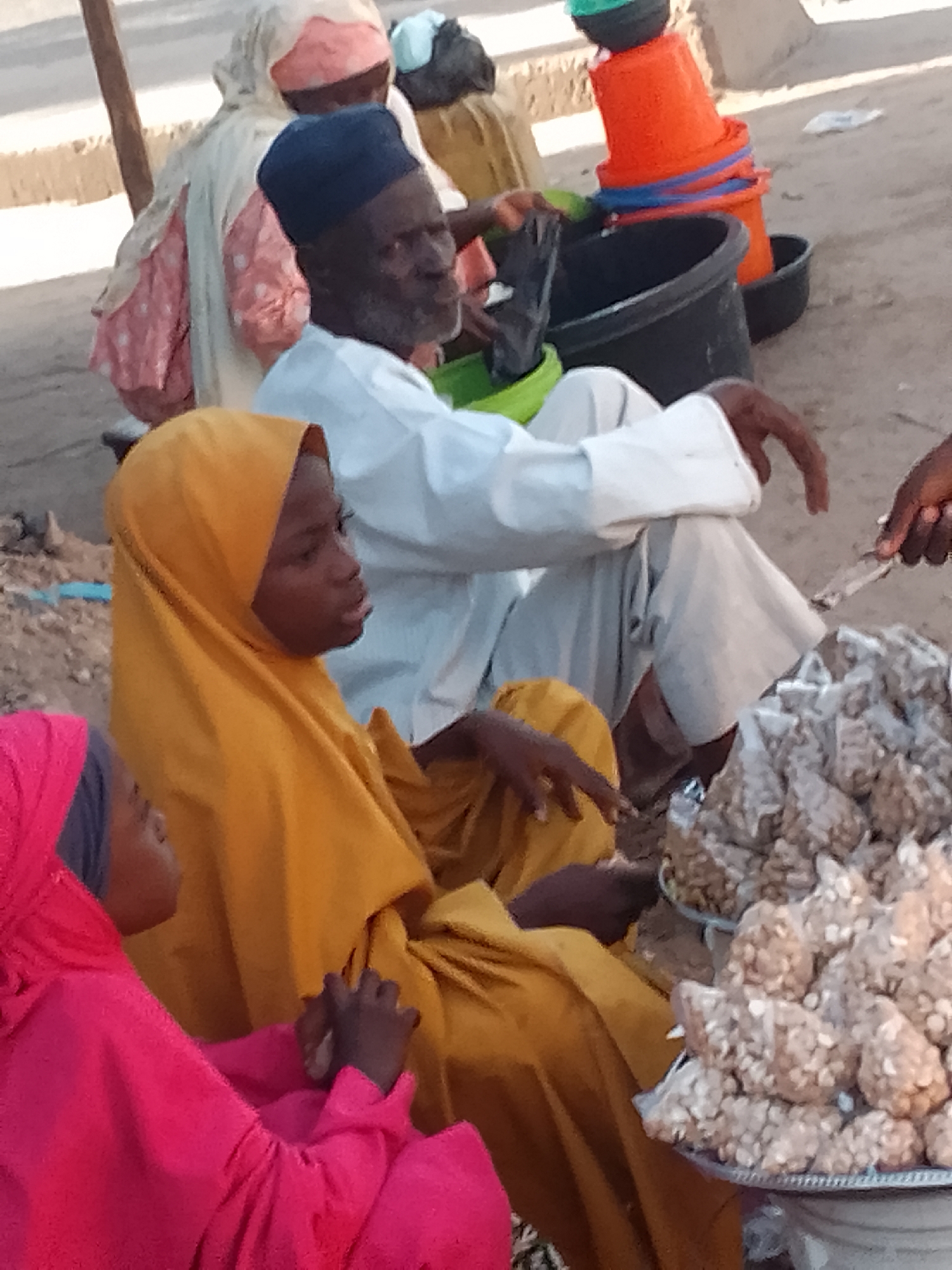 The image depicts a scene in Pantame, Nigeria. In the foreground, two young girls are visible. One girl, wearing a bright pink hijab, is facing left. Next to her, another girl wears a yellow hijab and is looking to the right. An elderly man, wearing a dark blue cap and a white robe, is seated and looking to the right. In the background, several individuals are partially visible, some wearing traditional clothing. Various objects are present, including stacked orange and blue buckets, and large black buckets, possibly for water or goods. A large pile of what appears to be nuts or seeds is displayed on a metal tray in the foreground, with a hand reaching towards it. The ground is sandy and dusty, suggesting an outdoor market or public space.