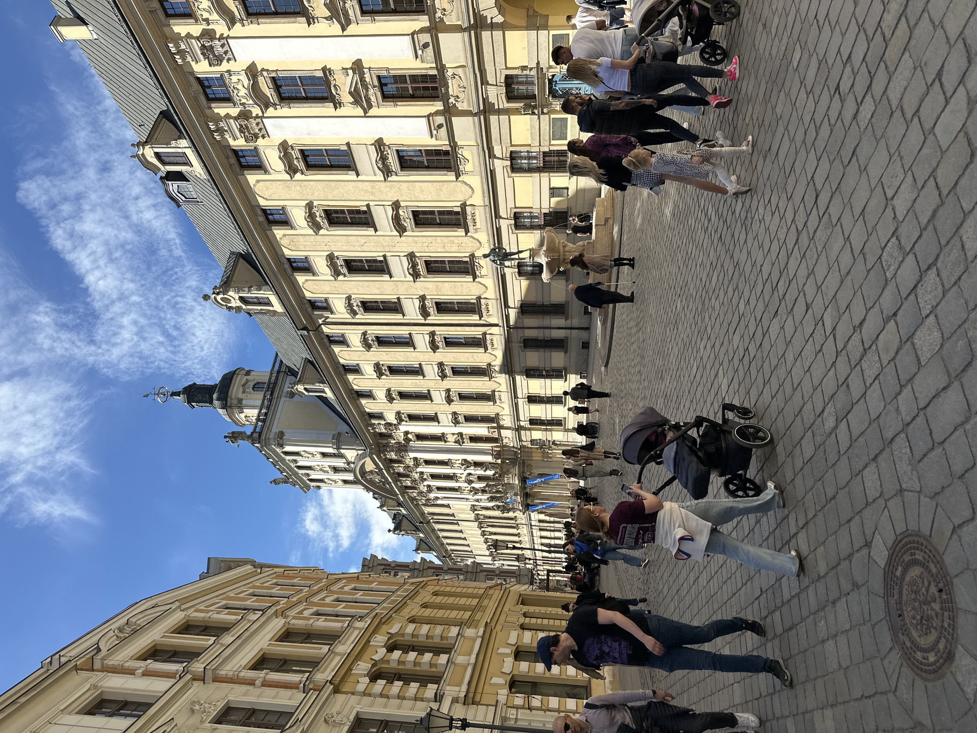 The image captures a street view in Wrocław, Poland. The scene is populated with pedestrians, including individuals and groups, some pushing strollers. Notable buildings are visible, showcasing European architectural styles with detailed window designs and ornamentation. The sky is a clear blue with light clouds, providing a backdrop to the buildings. The ground is paved with cobblestones, showing signs of wear. The lighting is bright, suggesting a sunny day.