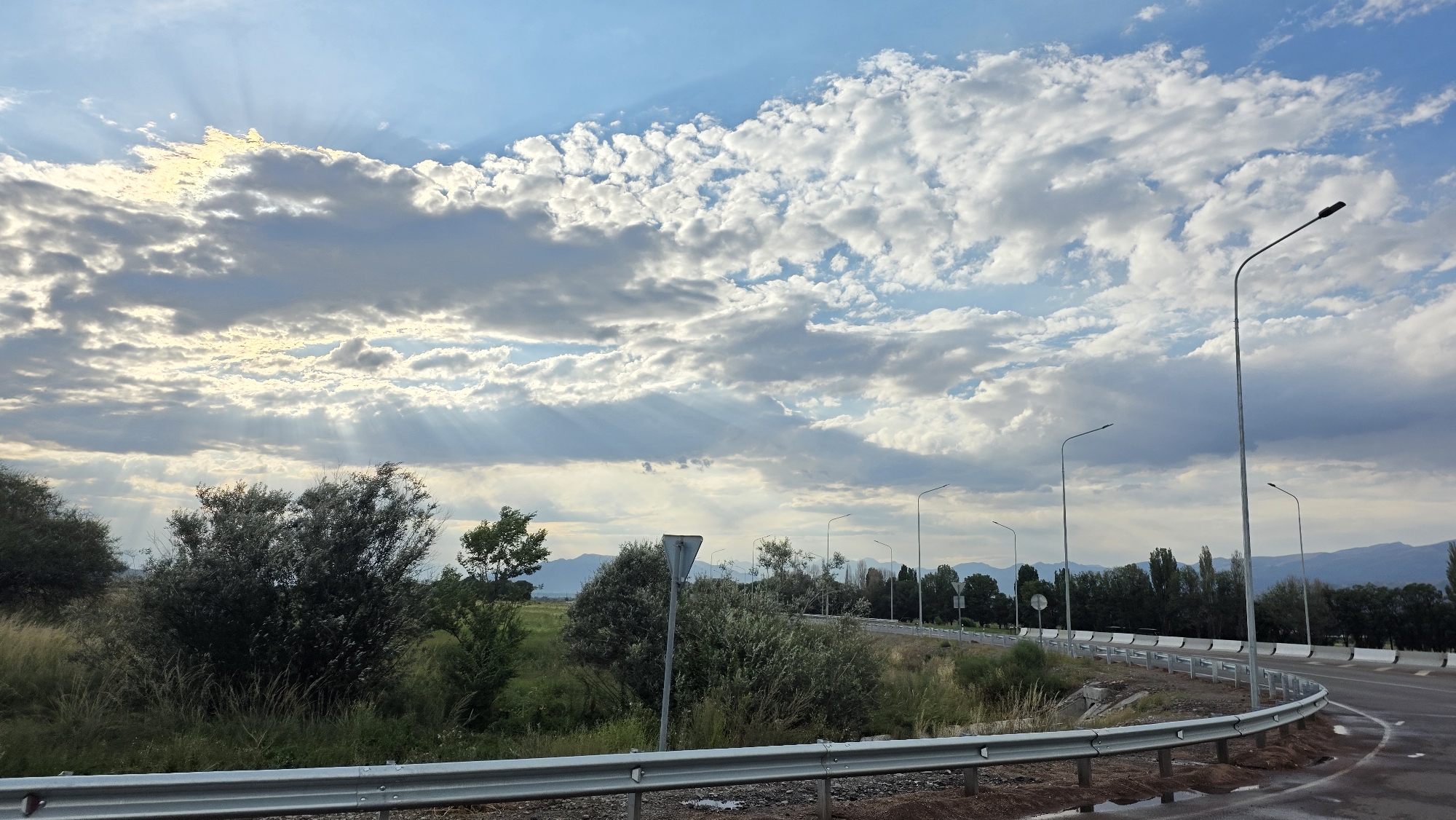 This image captures a scenic view of a road with a guardrail in the city of Kochkor, Kyrgyzstan. The sky is a vibrant blue with scattered clouds, and sun rays are breaking through, creating a dramatic effect. Several streetlights line the side of the road, which curves to the right, disappearing into the distance. To the left of the road, there is lush green vegetation, including trees and tall grasses, with a few bushes. In the background, a line of trees stretches across the horizon, and beyond them, mountains are visible under the cloudy sky. The road appears to be wet, suggesting recent rain. There are no people or vehicles visible in the image.