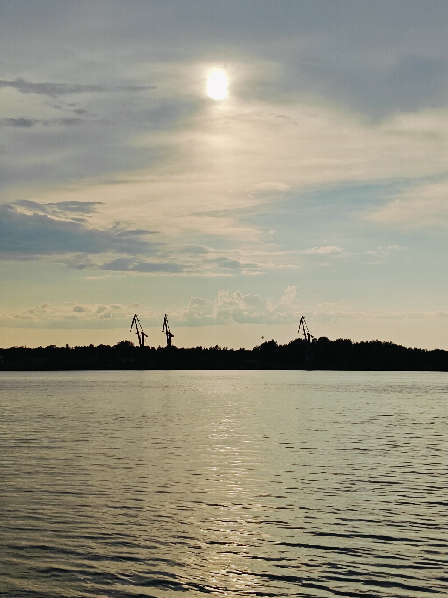 This image depicts a wide expanse of water, likely a river or bay, with a silhouetted treeline and industrial cranes along the distant shore. The sun is partially obscured by clouds, casting a bright glare on the water's surface, which is rippled by gentle waves. The sky is a mix of grey and pale blue clouds, with some wispy formations present. The overall scene suggests a waterfront industrial area, possibly during a cloudy sunset or sunrise. The industrial cranes imply a port or shipbuilding facility. Given the context of Chaikovsky, Russia, this likely represents a view of the Kama River.