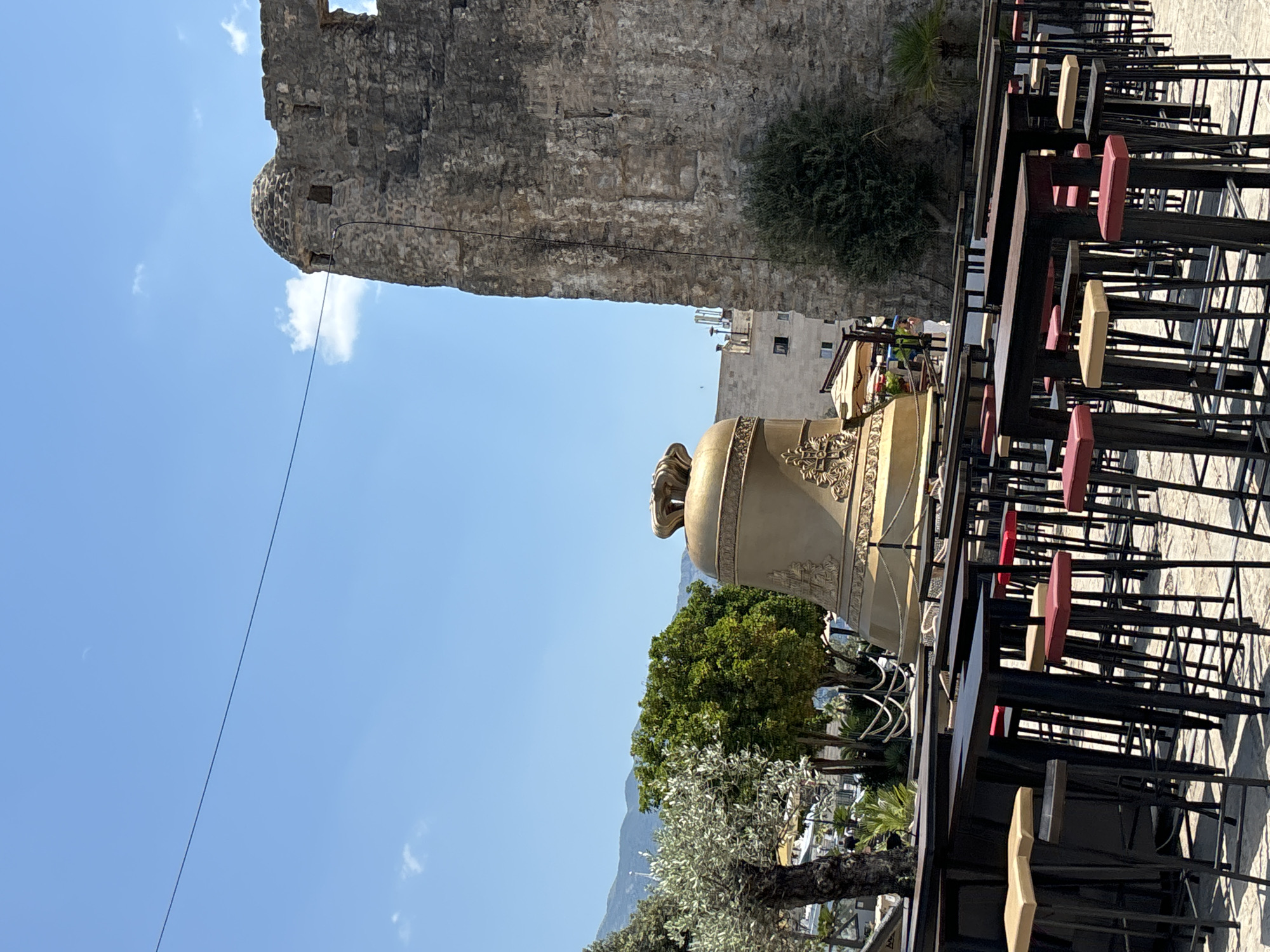 The image depicts an outdoor cafe setting with a view of ancient stone architecture and a bright blue sky with scattered clouds. In the foreground, rows of empty cafe tables and chairs are visible, some with colorful cushions. A large, ornate golden bell-like object stands prominently in the middle ground. Behind this, a segment of a rough stone wall, likely part of a historic structure, rises vertically. Further in the background, lush green trees and distant mountains are visible under the clear sky. The scene suggests a sunny day, possibly late morning or early afternoon, given the bright lighting and the presence of the moon in the sky. The overall impression is one of a serene, tourist-friendly location in Budva, Montenegro, offering a blend of history and leisure. No visible text is present in the image.