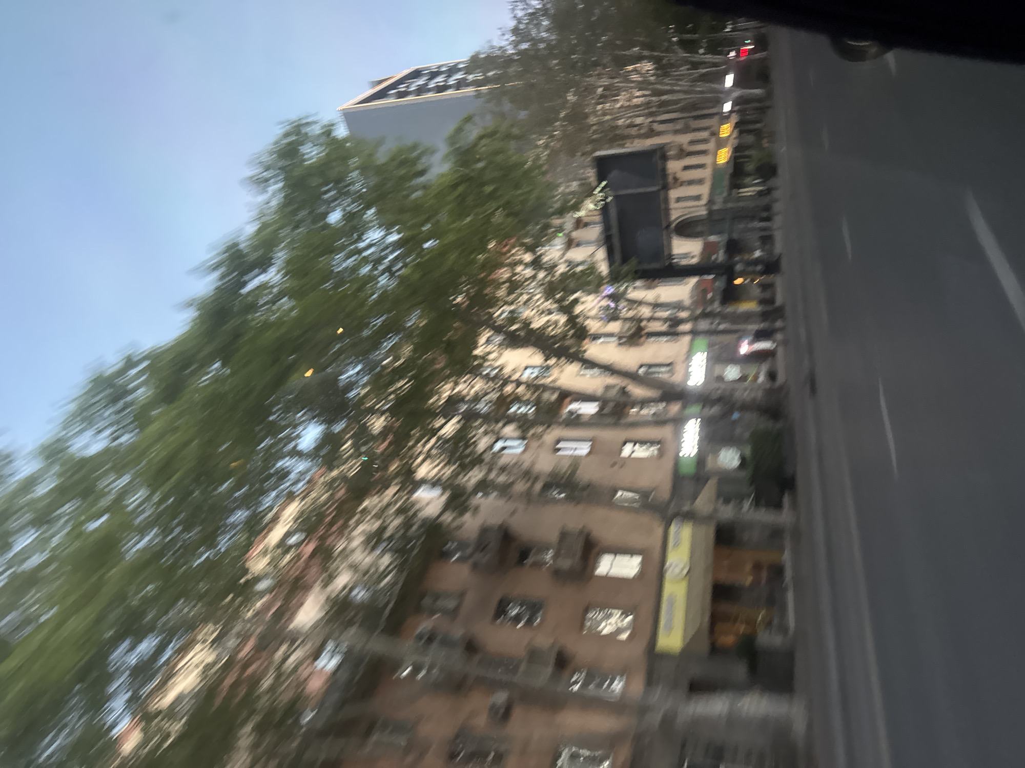 The image shows a street scene in Tbilisi, Georgia. A tall building, likely residential, is visible with multiple windows and balconies. A large, leafy tree obscures part of the building. A street sign is mounted on the building. To the right, a gray road stretches into the distance with painted white lines indicating lanes. A vehicle is likely on the road. The sky is blue and clear in the upper left corner of the image.