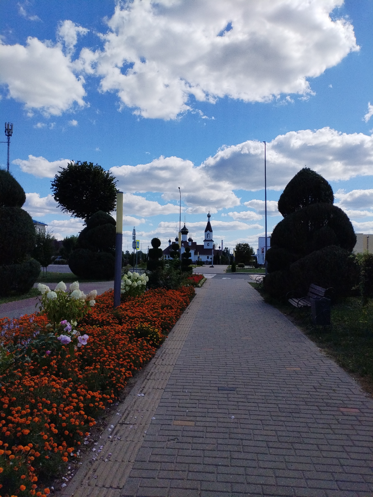 A paved walkway leads through a park area with manicured shrubs and flower beds. On the left, a vibrant flower bed is filled with orange marigolds and white hydrangeas. Further down the walkway, a wooden bench sits near a large, sculpted evergreen shrub. In the background, a white and gold church with multiple domes is visible. The sky above is blue with scattered white clouds. The scene appears to be in the city of Bialynichy, Belarus.