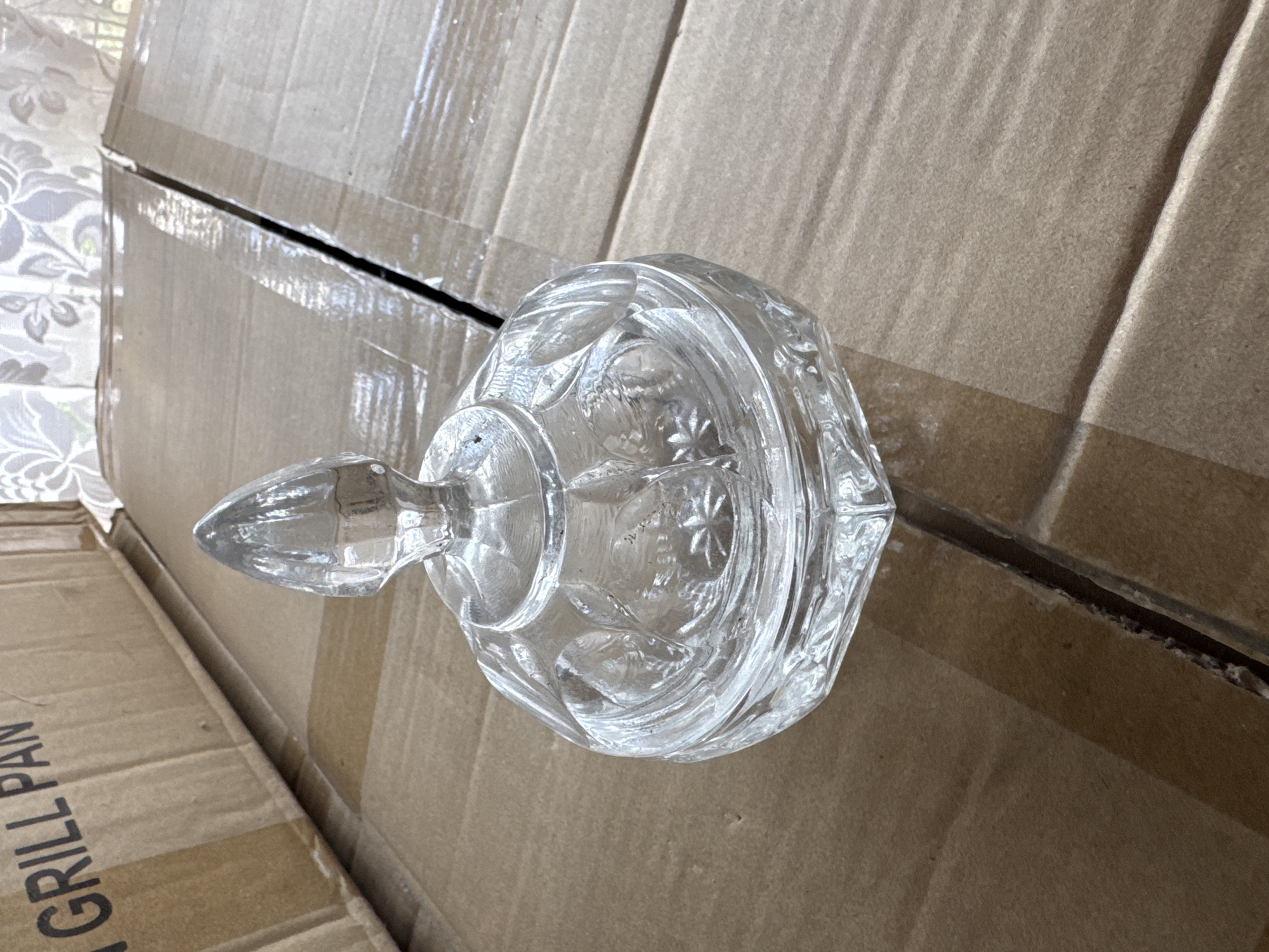 A clear glass candy dish with a lid sits on top of cardboard boxes. The lid has a pointed finial. The dish has a hexagonal base and is decorated with faceted patterns and starbursts. The boxes are brown and appear to be shipping boxes. The background shows a light-colored curtain with a floral pattern. The scene is indoors, possibly in a storage area or a home. The city of Rozdilna, Ukraine is not depicted in this image.