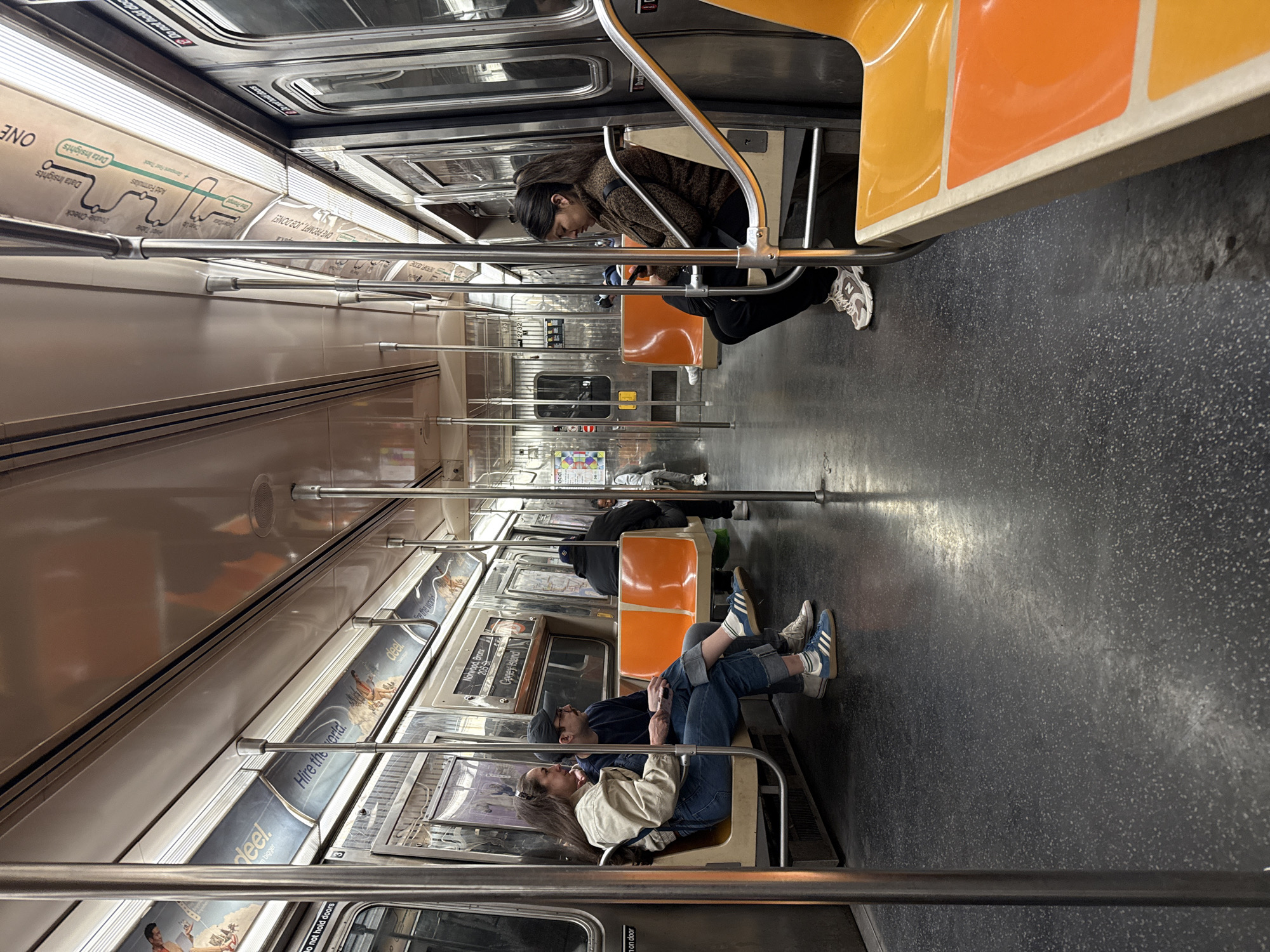 The image depicts the interior of a New York City subway car. Several passengers are visible: a woman is seated, looking at her phone; a couple is intertwined on a seat; and another person is seated further down the car. Subway route information and advertisements adorn the walls above the seats. The advertisements are for "deel." with the slogan "Hire the world." Other signs say, "Do not hold doors" and "One prompt, Job done!". The route information sign states, "Nohwood, Bronx 205-st."