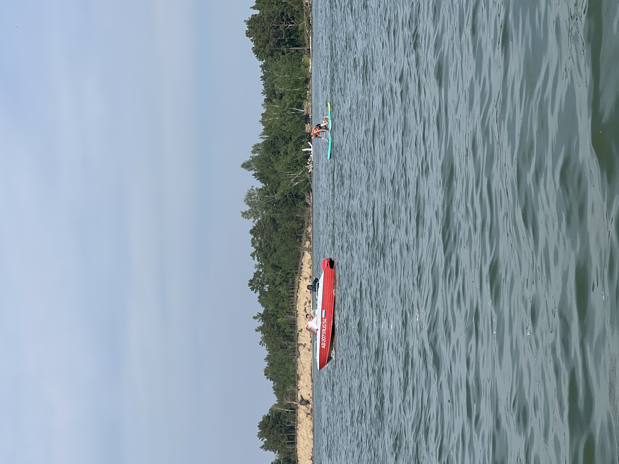 A bright red motorboat with the Russian flag and license plate "AB 2071RUS 54" is moving on the water. Two people are visible inside the boat. To the right, a person wearing a life vest is kneeling on a paddleboard. The background features a wooded shoreline with a sandy bluff. The scene appears to be a lake or large body of water in a natural environment, possibly near Borovoe, Russia, given the license plate. The water is rippled, indicating a light breeze. The sky is overcast with pale blue and white clouds.
