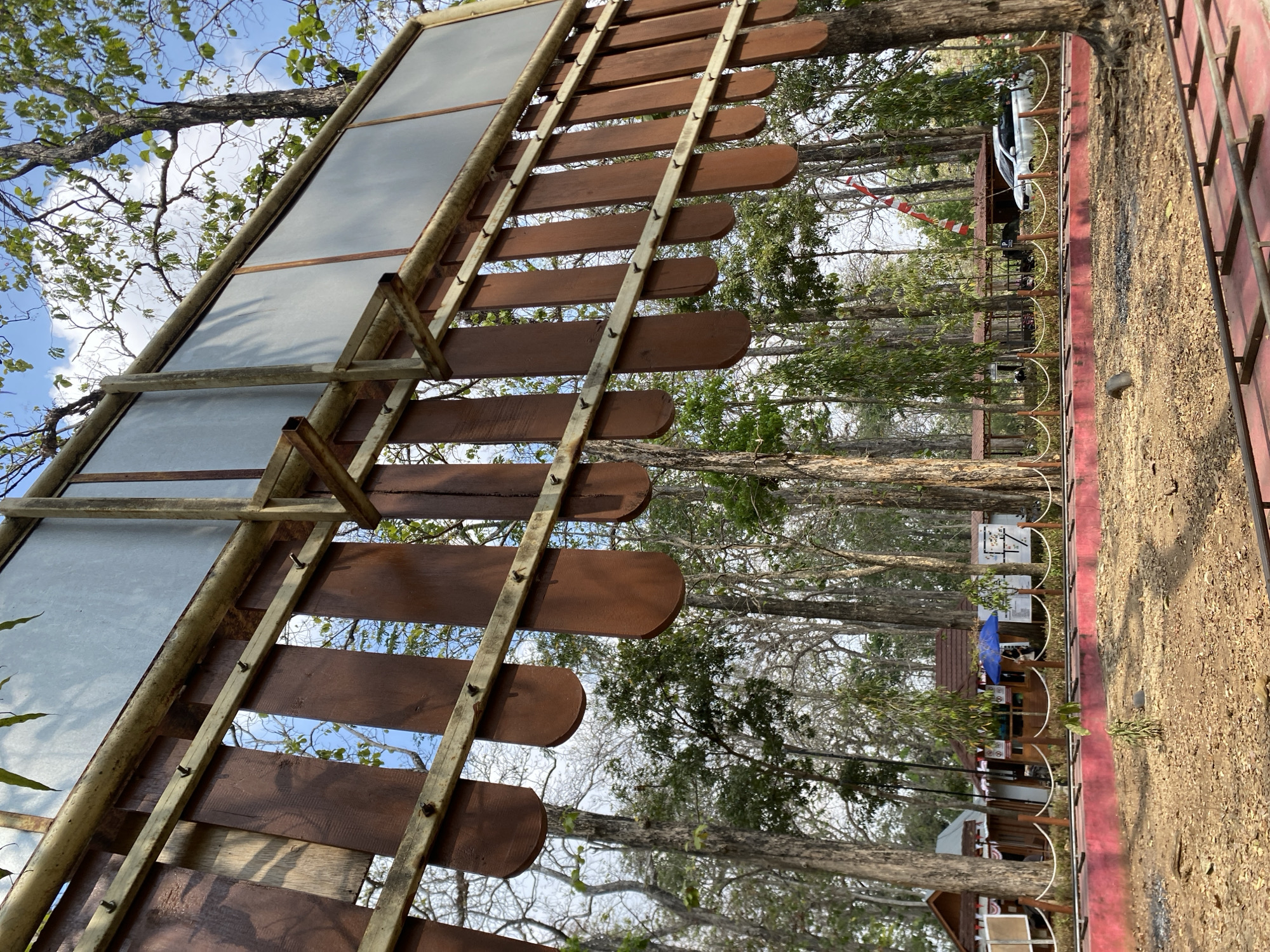 A low-angle, full shot captures a giant wooden xylophone-like structure set against a backdrop of tall trees and a bright blue sky with scattered clouds. The xylophone has wooden slats, painted dark brown and rounded at the bottom, suspended by metal bars. The frame and structural elements of the xylophone appear to be made of weathered metal. In the background, there are several small wooden structures, possibly stalls or pavilions, with red roofs. A blue umbrella is visible near one of the structures, and a white car is parked in the distance. A red-painted fence with rope railings separates the foreground from the background structures. The ground in the foreground is covered with dry leaves and dirt, with some metal tracks visible. This scene is located in Ketileng, Indonesia, which is known for its natural attractions and tourist spots.