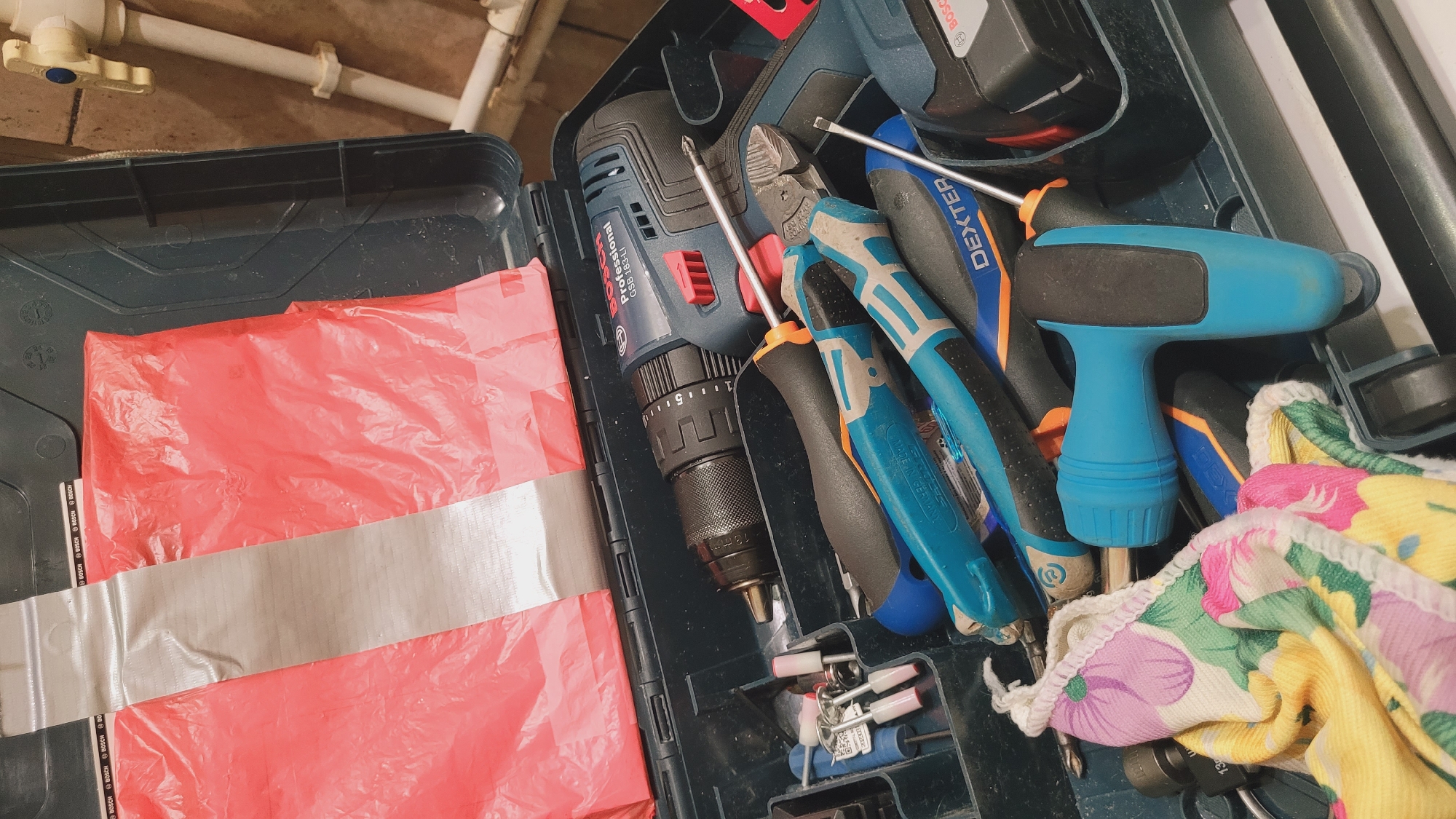 The image shows an open dark blue plastic toolbox filled with various tools. A red plastic bag, partially covered by silver tape, is placed on top of some of the tools. Prominent tools include a blue and black Bosch cordless drill, a pair of blue and black handled pliers, a blue handled screwdriver with a T-shaped grip, and a screwdriver with an orange and black handle. Other assorted bits and pieces are also visible within the toolbox. The background suggests an indoor environment, possibly a workshop or utility area, with plumbing pipes and a light-colored tiled wall visible in the upper right portion of the image. The lighting is dim and overhead.