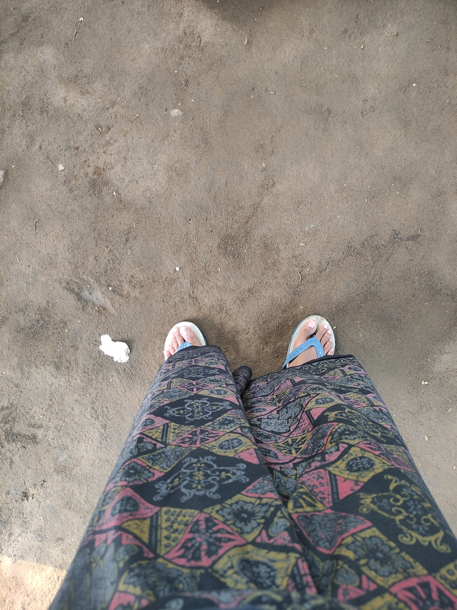 A person's legs and feet are shown from a top-down perspective, standing on a dirt ground. They are wearing a pair of white and blue flip-flops and a patterned skirt with black, pink, and yellow designs. A crumpled piece of white paper is visible on the ground to the left of the person's feet. The ground appears to be a dusty, hard-packed dirt surface with small pebbles and debris scattered across it. The lighting suggests it is daytime. This scene is likely in the city of Tiudan, Indonesia.