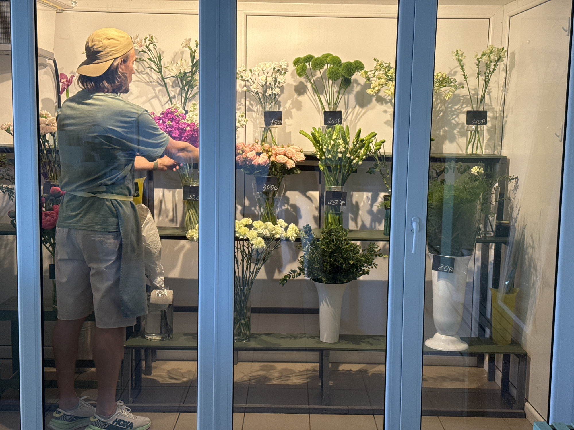 A man wearing a khaki baseball cap, a green t-shirt, and khaki shorts stands inside a flower shop, seen through the glass storefront. He faces the flower display, which features various arrangements in vases with price tags in Russian currency. The shop appears to be in Izhevsk, Russia, based on the Cyrillic alphabet on the price tags. The man is arranging flowers, potentially preparing them for sale. The shop has a minimalist aesthetic with metal shelving and a white interior. The exterior is a modern facade with clean lines.