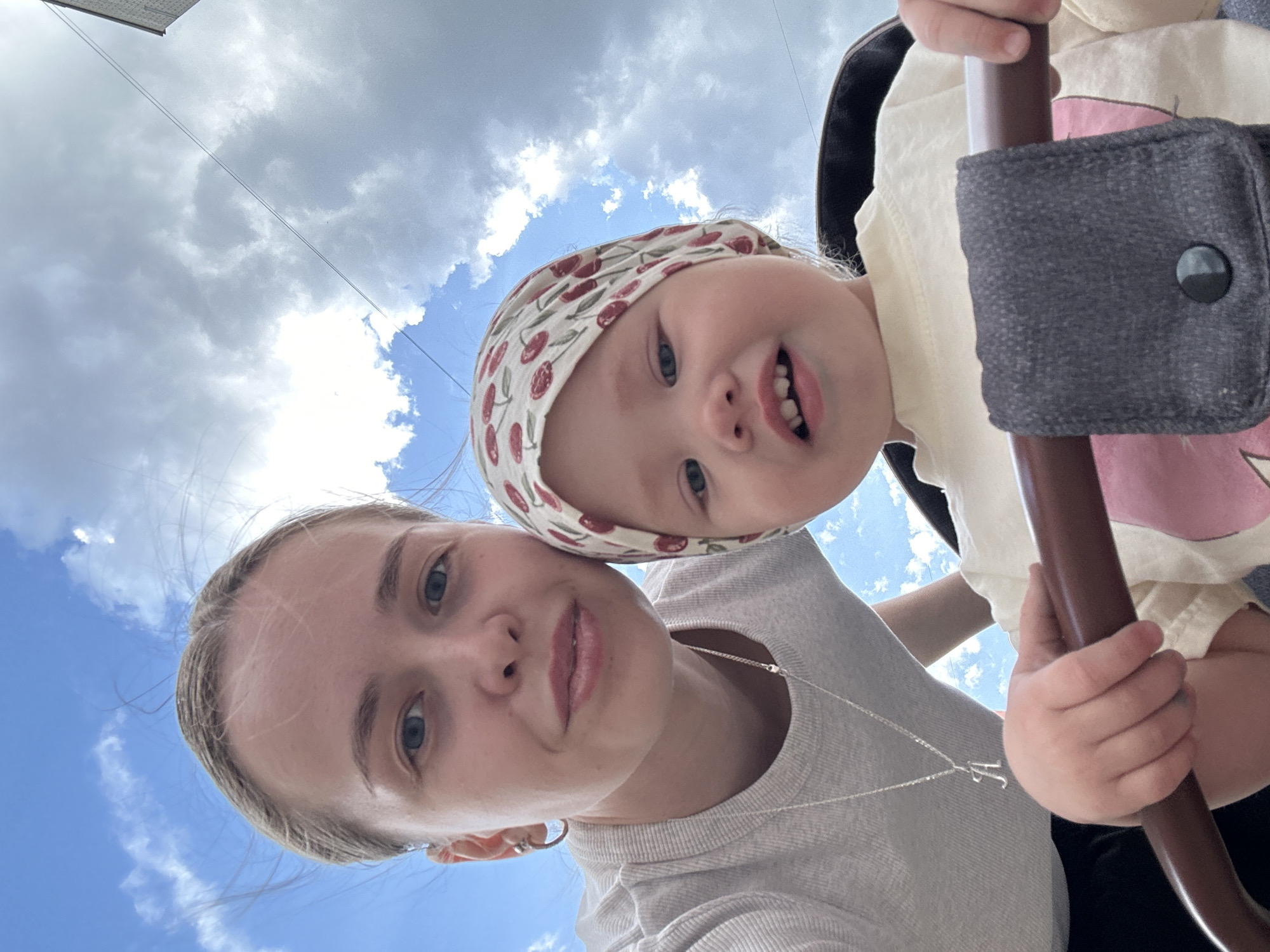 A selfie image shows a young woman and a baby. The woman has light skin and blonde hair pulled back. She wears a light-colored shirt and a silver necklace with a letter pendant. The baby is fair-skinned and wears a hat with a cherry pattern. The background shows a blue sky with scattered clouds. They are likely outdoors, possibly in a park, seated within some kind of buggy or carrier. No specific details indicate the location as Chaikovsky, Russia.