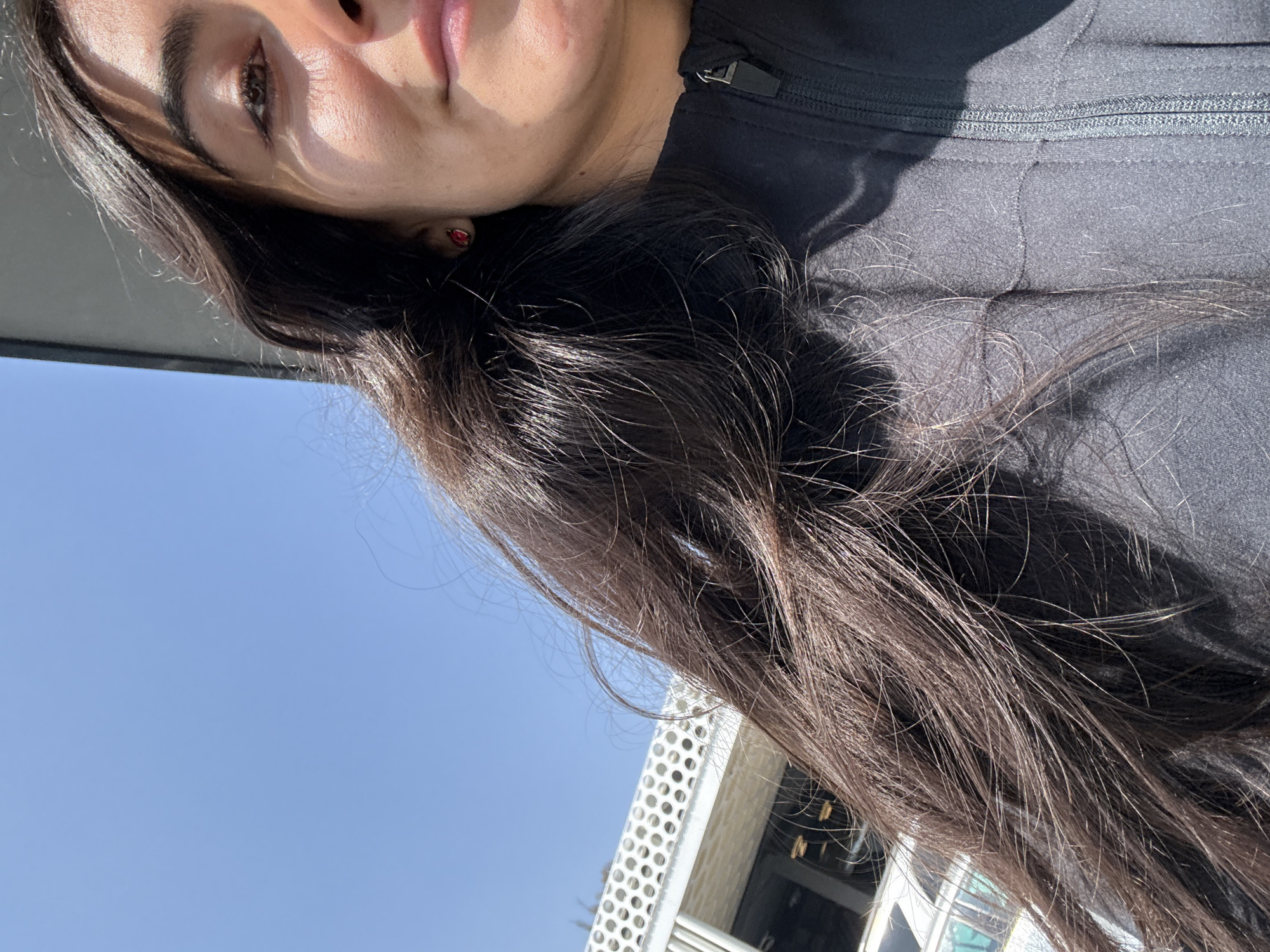 A close-up, outdoor shot depicts a female individual partialy visible from the right side of her face, including her right eye and nose, to the top of her head. Her long, dark brown hair cascades across the left and central portions of the frame. She wears a red stud earring in her right earlobe and a dark, possibly black, zippered jacket. Strong sunlight illuminates the scene, casting shadows across her face and hair. In the background, to the upper left, a portion of the Museo Internacional del Barroco in Puebla, Mexico, is visible, identifiable by its distinctive white, perforated facade featuring circular patterns against a clear blue sky.