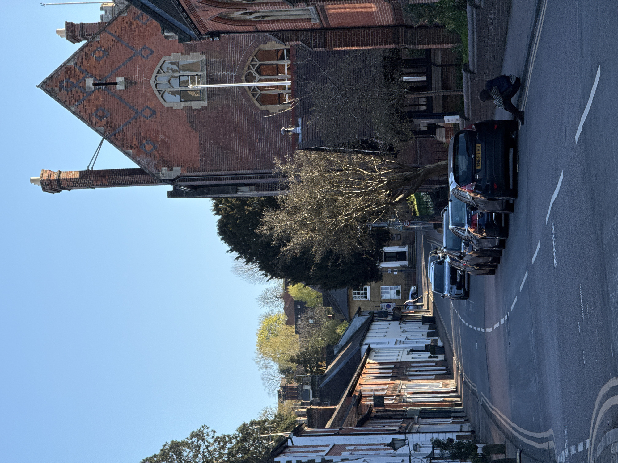 A street view in Dacorum, United Kingdom. The road has cars parked along the right side. A person bends over near a parked black car. Buildings with varying architectural styles line the street, including a brick building with a distinctive roof pattern and arched window. Trees are interspersed among the buildings, and the sky is clear.