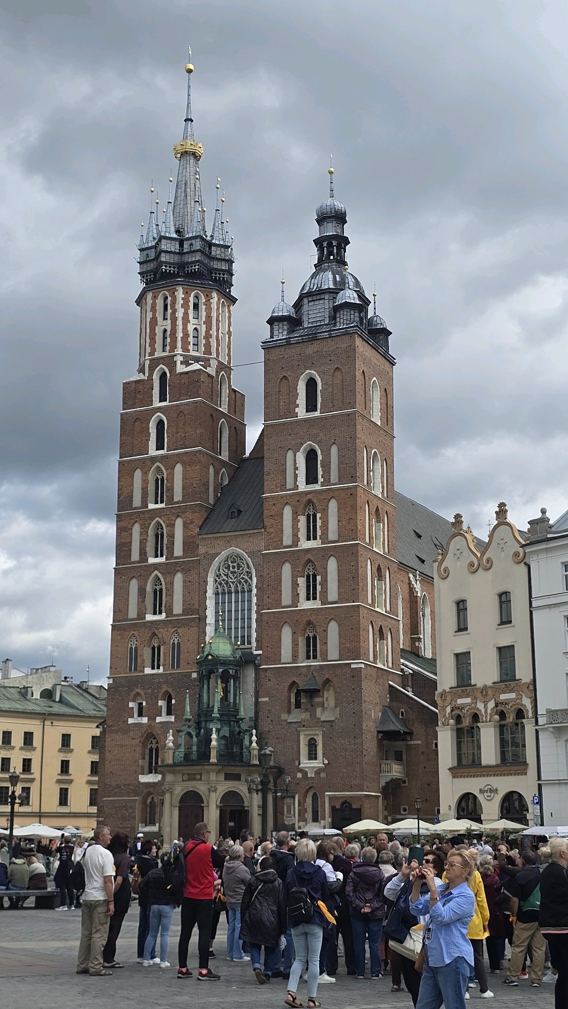 The image shows a wide view of St. Mary's Basilica in Krakow, Poland, a prominent landmark in the city's Main Market Square. The basilica's iconic brick towers, with their distinct Gothic architecture, dominate the frame. The left tower is taller and features a spired roof with gold accents, while the right tower is shorter and capped with a dark, multi-tiered dome. In front of the basilica's entrance, a green-domed structure with intricate metalwork stands out.
A diverse crowd of people is gathered in the square, many of whom appear to be tourists. Some are standing, observing the basilica, while others are engaged in taking photographs. A woman in the foreground, wearing a blue shirt and jeans, is actively taking a picture with her phone. Several people are dressed casually, suggesting a daytime excursion. In the background, other buildings with traditional European architecture are visible, along with streetlights and some market stalls with umbrellas. The sky is overcast with gray clouds, creating a subdued lighting condition.