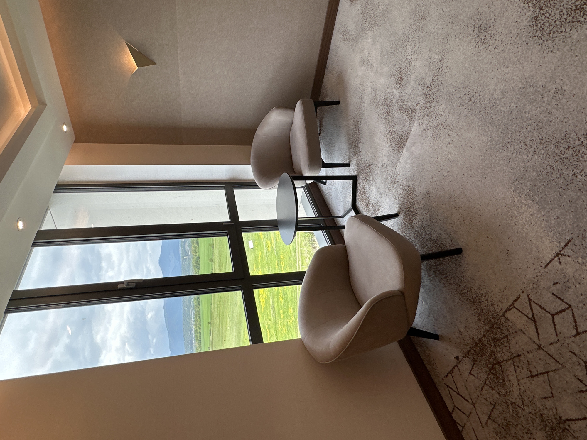 This image depicts a modern, minimalist room interior with a panoramic window view of an outdoor landscape, located in Kazanlak, Bulgaria. The room is furnished with two contemporary, light beige upholstered armchairs, each with black legs, positioned to face the large window. A small, round black side table sits between the chairs.
The floor is covered with a textured, light-colored carpet featuring a subtle, abstract geometric pattern in darker brown or grey tones. The walls and ceiling are a uniform, light, neutral color. The ceiling incorporates modern lighting elements, including recessed spotlights and a distinctive triangular wall sconce that directs light upwards.
Through the expansive, multi-pane window, a vibrant daytime scene unfolds. Lush green fields, interspersed with patches of bright yellow vegetation (possibly flowering crops), stretch towards a backdrop of distant, undulating mountains or hills. The sky is overcast with soft, white clouds, indicating a cloudy or partly cloudy day with diffused natural light illuminating both the interior and exterior.
The scene suggests a quiet, contemplative setting designed for relaxation, offering a direct connection to nature without any visible human activity or text.
