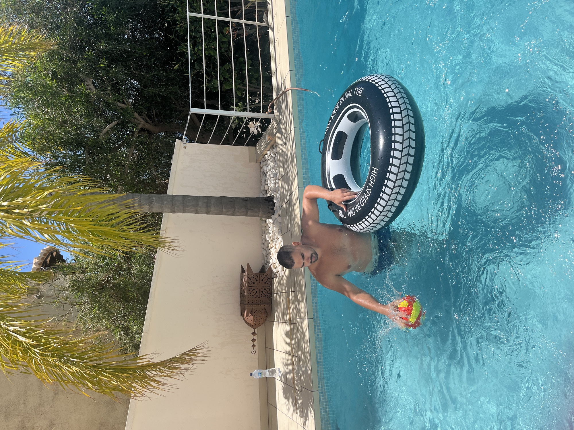 A medium shot captures a man in a swimming pool, potentially in a residential area of El Poble Nou de Benitatxell/Benitachell, Spain. He is partially submerged, wearing what appears to be blue swimming trunks, and is holding a brightly colored ball in his right hand, creating a splash. The man is also using his left arm to hold onto a black and white inflatable tire.
To the left, the swimming pool is bordered by a cream-colored wall and tiled edge. Beside the wall are decorations, a water bottle, and white pebbles. Above the wall, foliage and trees are visible. A palm tree with yellow leaves is positioned to the left, partially obscuring the scene. Sunlight illuminates the scene.