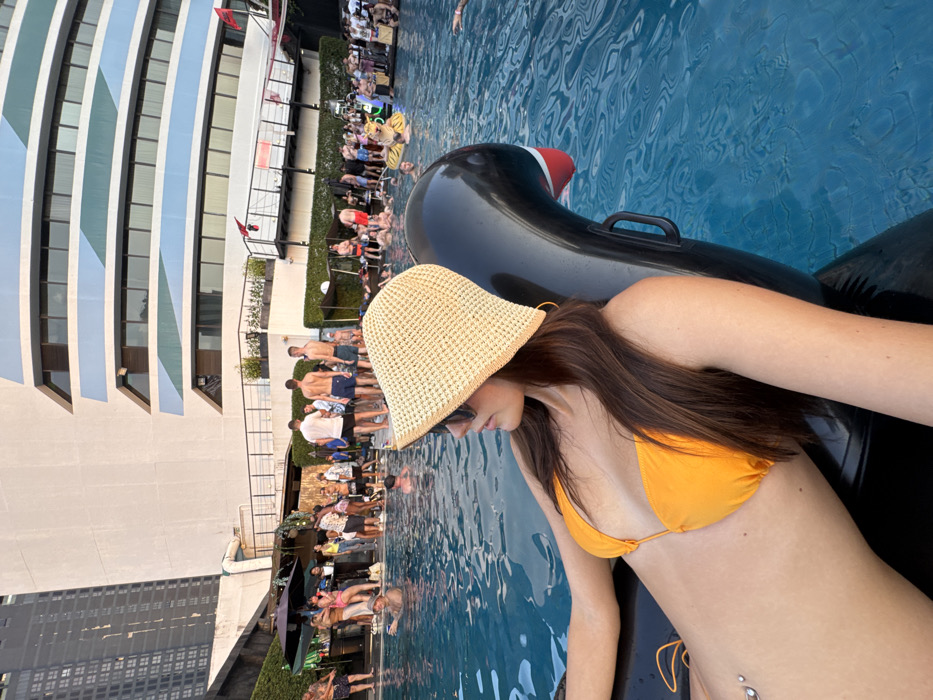 The image depicts an outdoor pool party scene. In the foreground, a female individual with long brown hair, wearing an orange bikini top, a straw bucket hat, and sunglasses, is partially submerged while reclining on a black inflatable swan float in a clear blue swimming pool. A navel piercing is visible on her abdomen.
The midground and background show numerous other individuals, both male and female, in various states of activity; some are wading in the pool, while others are standing on the surrounding poolside deck. The deck area includes a bar, several umbrellas, and a gathering of people. An additional yellow, tiger-striped inflatable float is visible in the pool further back.
Dominating the background is a multi-story, modern hotel building with a light-colored facade, distinguished by prominent horizontal blue and grey striped bands. A taller, darker skyscraper is partially visible to the left of the hotel. This scene is located at the SO/ Bangkok hotel's pool area, in Bangkok, Thailand.