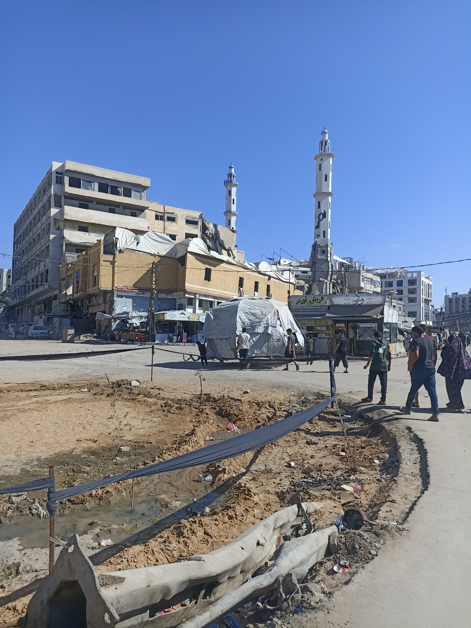 The image depicts a street scene in what appears to be a conflict-affected area, possibly Gaza. In the foreground, a trench has been dug, with debris and some vegetation present. A dark fabric is draped across the trench. A concrete structure, possibly a damaged barrier, lies in the lower part of the image.
In the mid-ground, several people are walking on a paved road. Behind them, a large, damaged building dominates the background, with sections appearing to be under construction or repair, covered with white tarps. Two minarets of a mosque are visible rising above the damaged structures, one of which appears to have sustained damage as well. A tent-like structure made of white fabric is also present. Several shops with signs are visible.
The sky is clear and blue, suggesting a sunny day, likely mid-morning or early afternoon given the shadows. The overall impression of the scene is one of ongoing reconstruction or recovery amidst evident destruction. Some of the visible text appears to be Arabic signage above the shops.