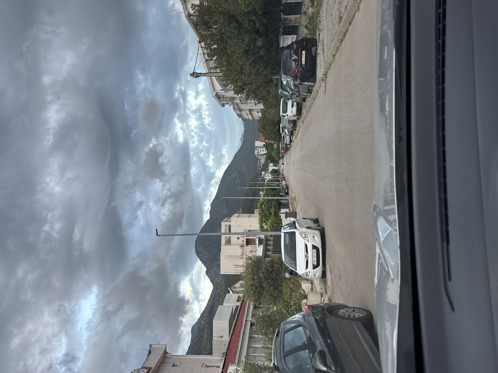 The image depicts a street scene in Sutomore, Montenegro, viewed from inside a vehicle. The street is lined with parked cars on either side, leading towards a distant mountain range under a cloudy sky. Buildings of varying heights and architectural styles are visible along the street. A lamp post is positioned on the left side of the road.