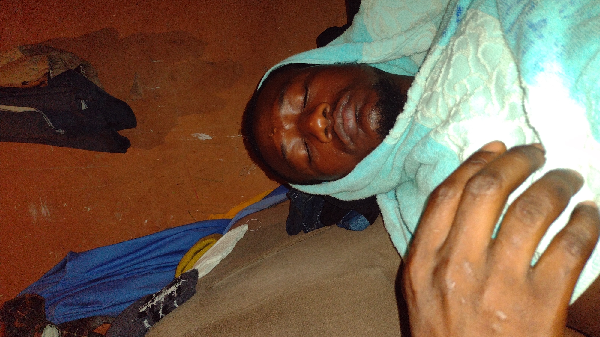 A close-up, overhead shot shows a Black man with his eyes closed, lying on his side. He is wearing a light blue hooded towel around his head and shoulders, the towel has a white embroidered pattern. His left hand is visible in the foreground, resting on the towel. He has a beard and is wearing a dark-colored shirt beneath the towel. The background is a textured, reddish-brown wall. To the left of the man, a brown cushioned surface is visible, along with a folded blue garment and other clothing items. The scene appears to be indoors.