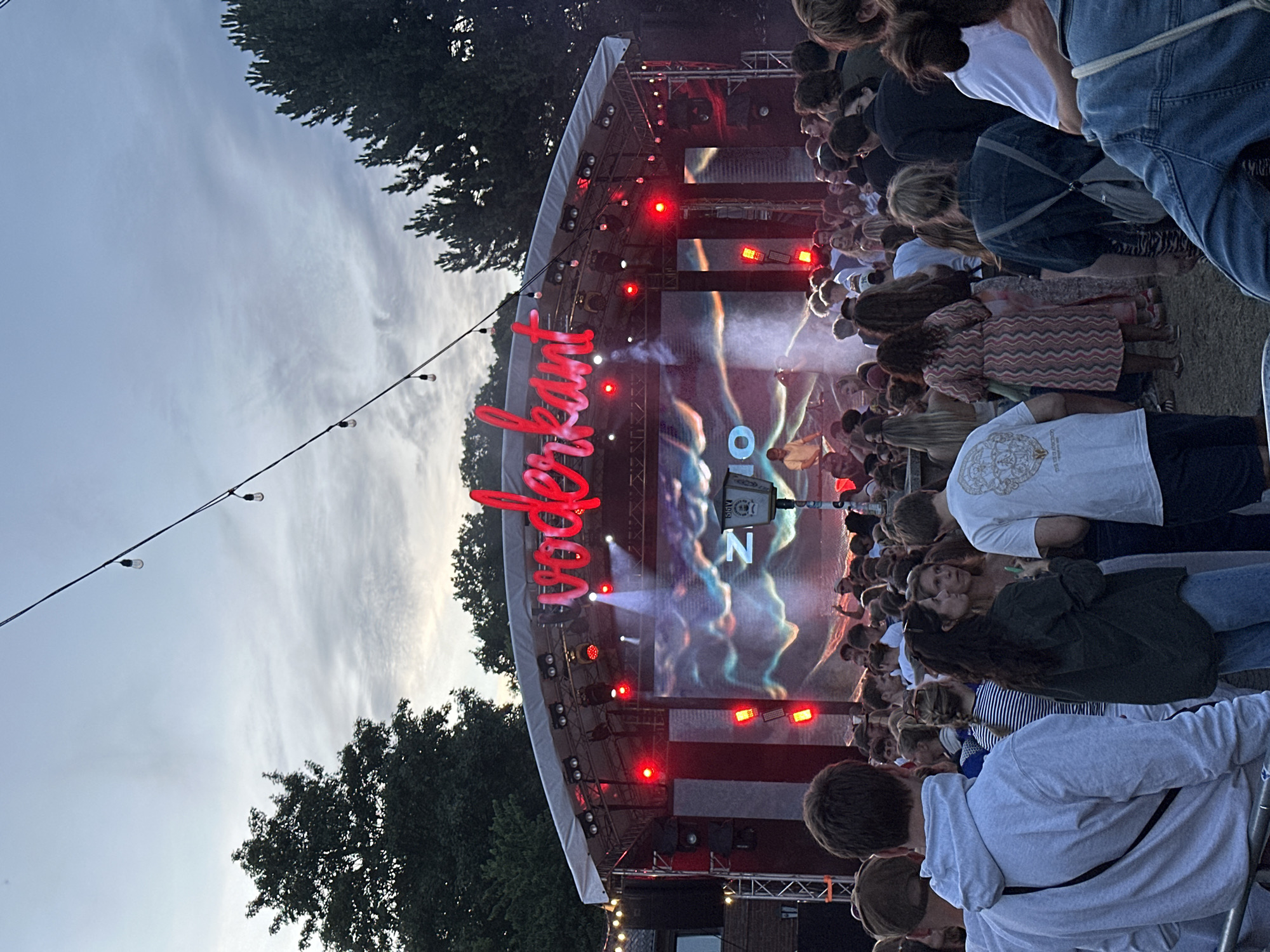 The image depicts an outdoor concert or festival scene. A large stage is set up, featuring a screen displaying abstract visuals. Red lights are prominent on the stage, illuminating the performance area. A large, red, stylized text banner with cursive lettering is positioned prominently on the stage, partially obscured by smoke or haze, but the word "ZODIAC" is clearly visible. Below the stage, a crowd of people are gathered, facing the stage and appearing to be enjoying the event. The overall atmosphere seems lively and festive. The time of day appears to be dusk or early evening, with the sky visible in the background showing a cloudy, twilight sky. Trees are visible on the left and right edges of the frame, suggesting the event is taking place in an outdoor, possibly park-like, setting. String lights are hung in the foreground, adding to the festive ambiance. The people in the crowd are dressed casually, in a mix of t-shirts, hoodies, and jeans. The scene suggests a communal gathering focused on entertainment, with a clear emphasis on the performance happening on stage. The presence of a stage, screen, lighting, and an audience all point to a live event. The clear visibility of the text "ZODIAC" on the stage is a notable detail.