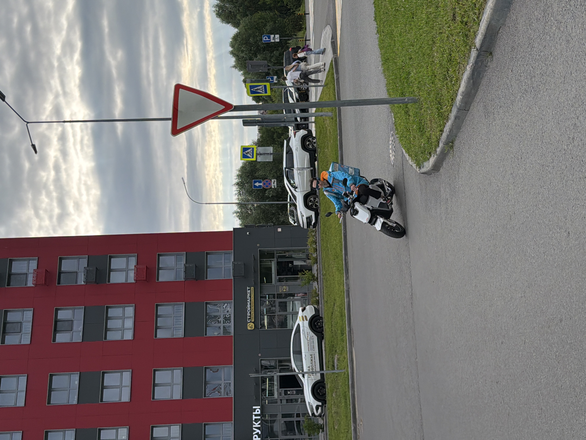 A person on a white scooter with a blue delivery box is driving down a paved road. The scooter has a license plate that is difficult to read. The person is wearing a blue jacket and an orange helmet. A white car with a black roof is parked on the side of the road. Another white car is partially visible in the background. A red building with many windows lines the left side of the road. A sign on the building reads "СТРОЙМАРКЕТ". A green grassy area is on the right side of the road. There are several traffic signs, including a yield sign and a pedestrian crossing sign. The sky is cloudy. The image appears to have been taken during the day.