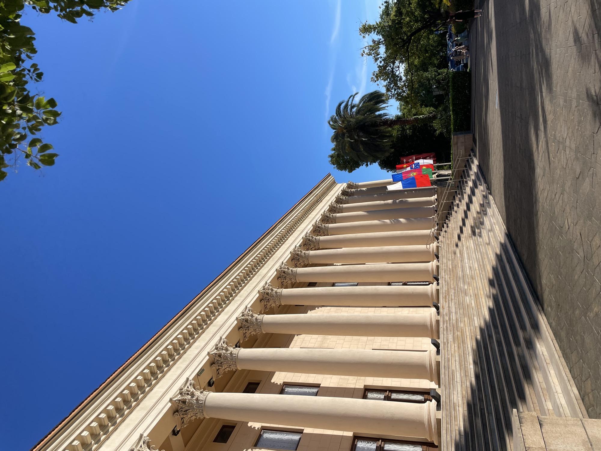 A wide-angle, low-angle shot captures the facade of a grand, neoclassical building on a sunny day in Sochi, Russia. The building, constructed from light-colored stone, is adorned with a row of tall, fluted Corinthian columns supporting an ornate entablature. Large, rectangular windows with white curtains are visible between the columns. A wide, stone staircase leads up to the entrance, its steps casting long shadows across the pavement.
In front of the building, a line of flags, including the Russian flag, are displayed. To the right of the staircase and flags, a lush palm tree stands tall amidst other greenery. Further in the background, several people are walking on a paved area, and cars are parked. The sky is a clear, vibrant blue with a few wispy white clouds. Lush green trees frame the right side of the image, their leaves catching the sunlight. The overall scene conveys a sense of grandeur and tranquility.