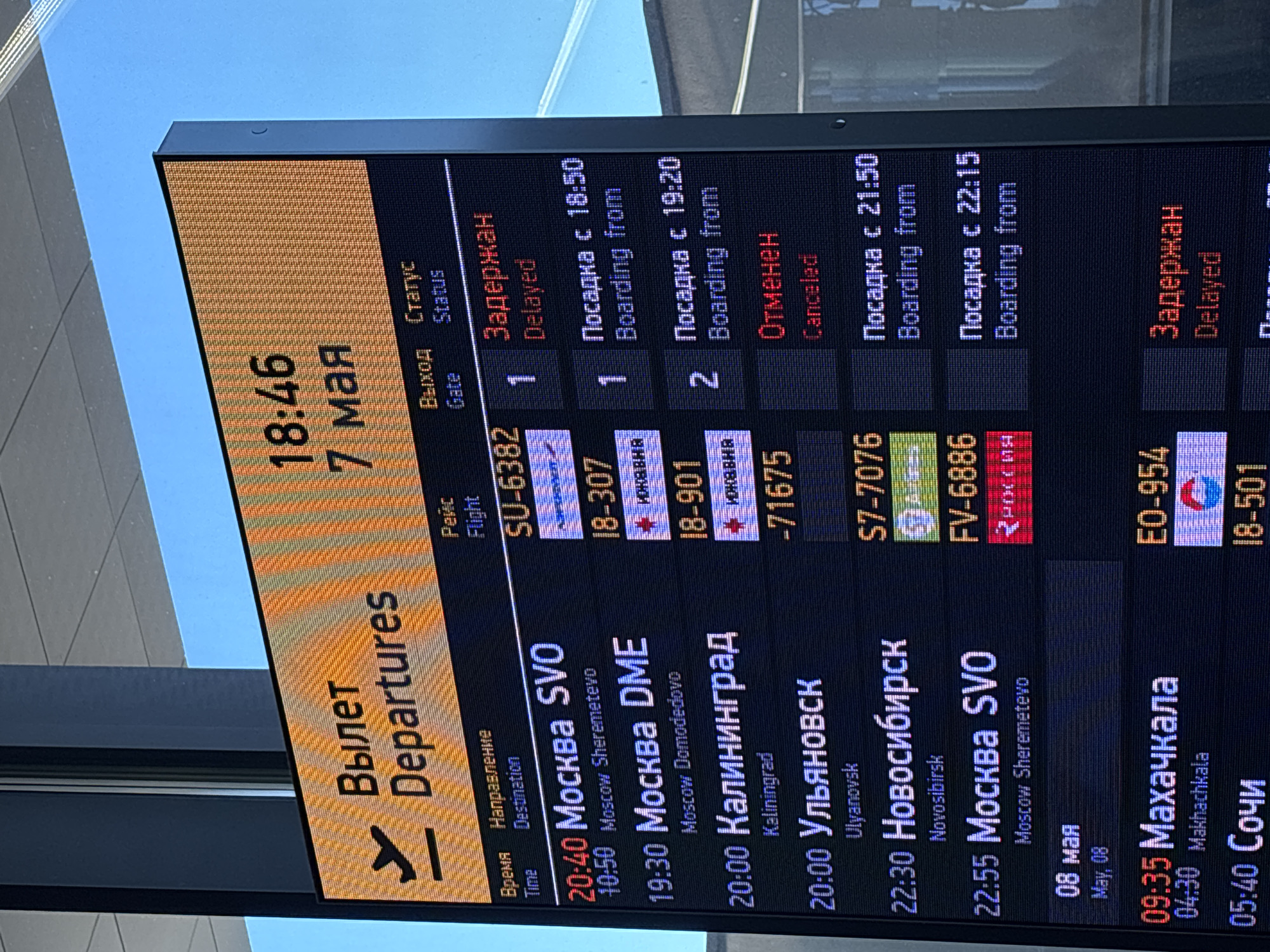 The image shows a departures board at an airport, displaying flight information. The board lists the time, destination, flight number, gate, and status of various flights. Some flights are delayed, one is canceled, and others are boarding. Destinations include Moscow (SVO and DME), Kaliningrad, Ulyanovsk, Novosibirsk, Makhachkala, and Sochi. The date shown on the board is May 7, with one flight listed for May 8. The location is not Новое Мартьяново; the flights listed depart from Moscow airports.