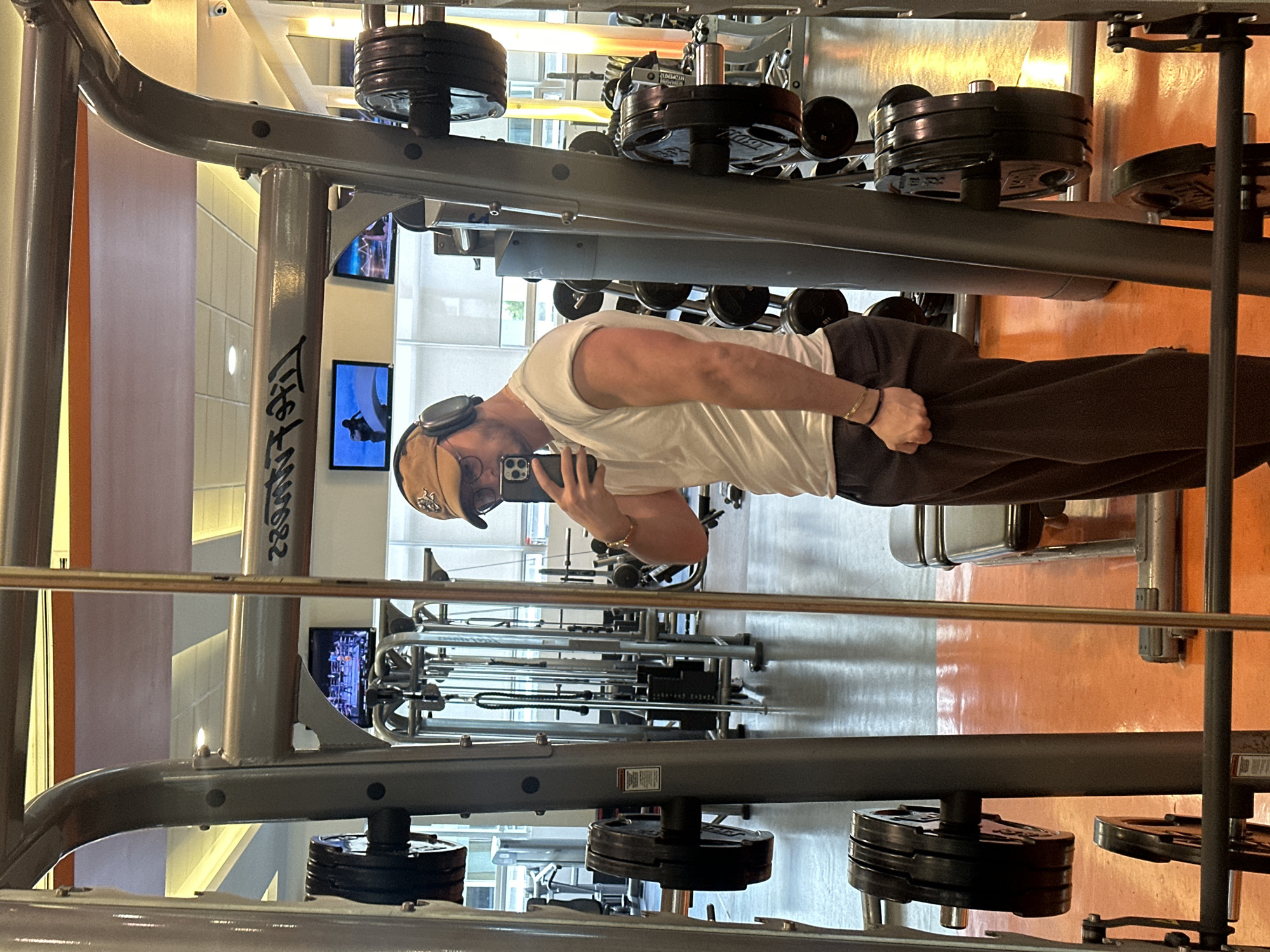 A person in a white t-shirt, brown pants, and a brown cap is standing in a gym, holding a smartphone and taking a picture. The gym has weightlifting equipment with stacked weights, and multiple workout machines are visible in the background. The environment has a light orange and beige color scheme, and television screens hang on the walls. The location is Mexico City, Mexico.