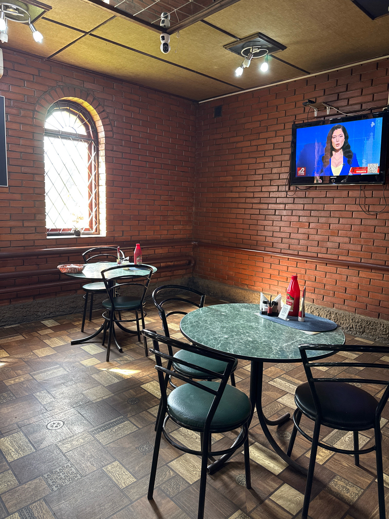 This image shows an interior view of a cafe or restaurant in Novyi Berezai, Russia. The walls are constructed of red brick, and the ceiling is paneled with yellow material, with a grid of metallic strips. A flat-screen television is mounted on the brick wall, displaying a news anchor. There are several tables and chairs in the room. Two round tables with green marble-patterned tops are visible, each with black metal chairs. On one table, there is a red bottle, napkins, and salt and pepper shakers. A large arched window with a diamond-patterned grate is present on the left side of the room, letting in natural light. The floor is covered with patterned linoleum tiles. There are two security cameras visible on the ceiling. The overall impression is of a simple, functional indoor dining space.