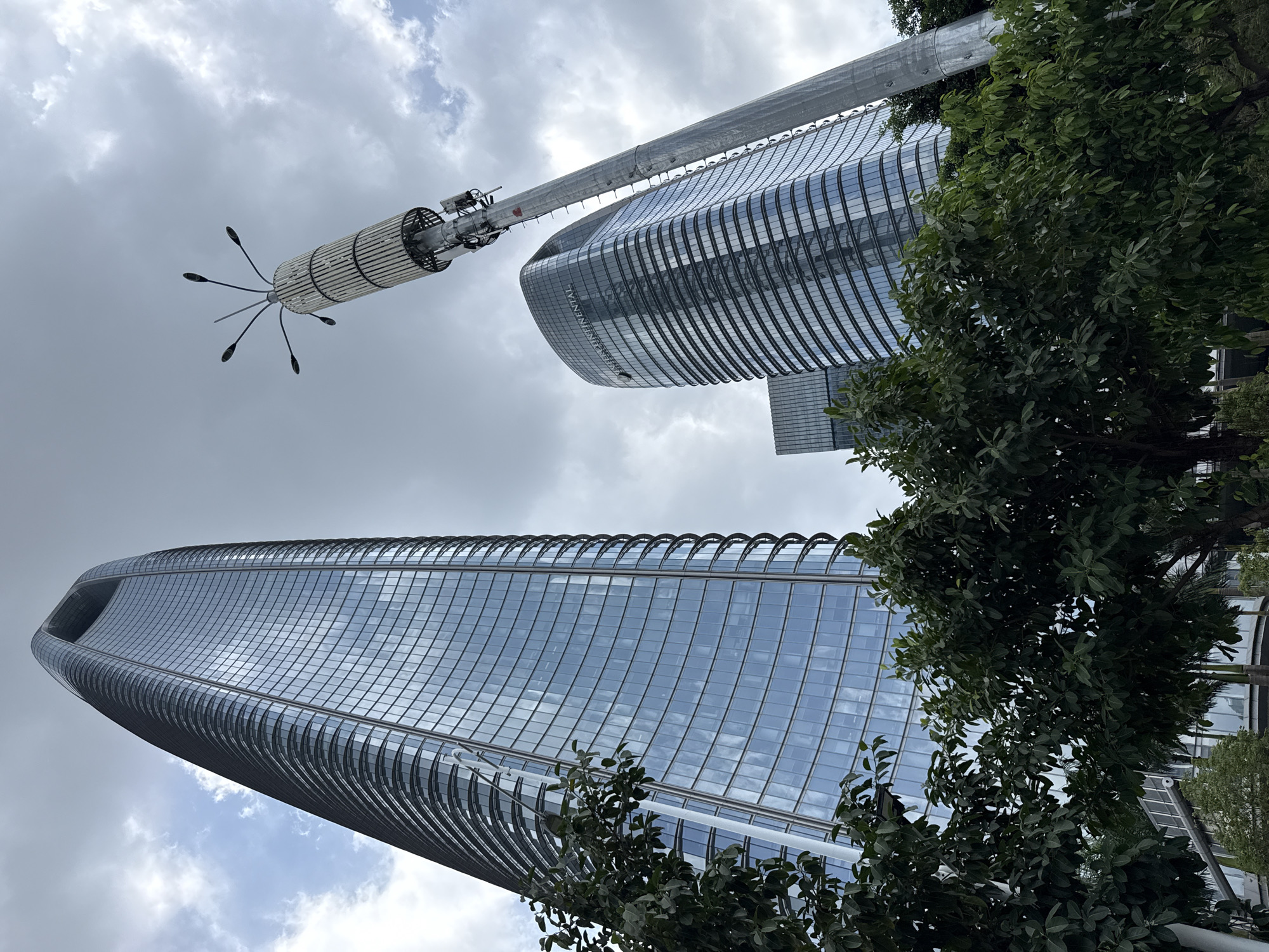 An eye-level, wide-angle, exterior shot captures a collection of modern skyscrapers and a cellular tower against a cloudy sky. The foreground features lush green trees. Dominating the left side of the frame is a tall, slender, glass-clad skyscraper with a distinctive curved top. To the right and slightly behind it, another curved skyscraper, identified by the "INTERCONTINENTAL" logo, stands prominently. A tall, metallic cellular tower with a cylindrical, ribbed section and an array of antennas at its apex is situated between the two buildings and leans into the sky. In the background, a third, less distinct skyscraper is visible. The scene is set in the Haizhu District, China.