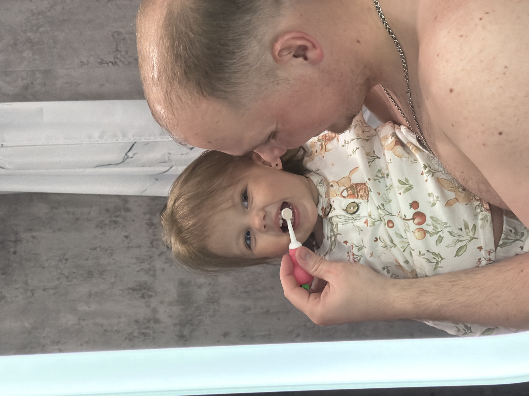 A man is brushing a child's teeth in a bathroom. The man is shirtless, balding, and wearing a silver chain. The child is wearing a patterned top with a bunny design. The bathroom has a gray wall and a white countertop. There is no indication of this scene being in Reshetnikovo, Russia.