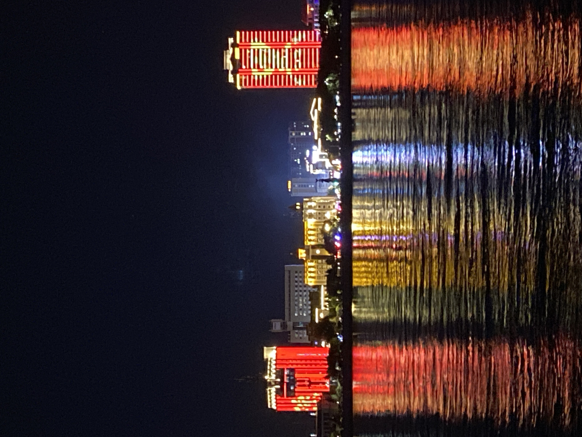 The image depicts a nighttime cityscape with a body of water in the foreground. Several buildings are illuminated with colorful lights, casting reflections onto the water. On the left, a tall, rectangular building is prominently lit with vibrant red lights, creating a striking visual. To its right, a series of lower buildings are also illuminated, with some featuring golden and yellow lights. Further to the right, another tall building stands out with a pattern of red lights. The water surface shows dynamic reflections of these lights, creating abstract patterns of red, yellow, and blue. The sky above is dark, indicating it is nighttime. The scene suggests a modern urban environment. This scene is of the Blagoveshchensk embankment in Russia, specifically showing the skyline across the Amur River, likely facing Heihe, China. There are no people visible in the image.