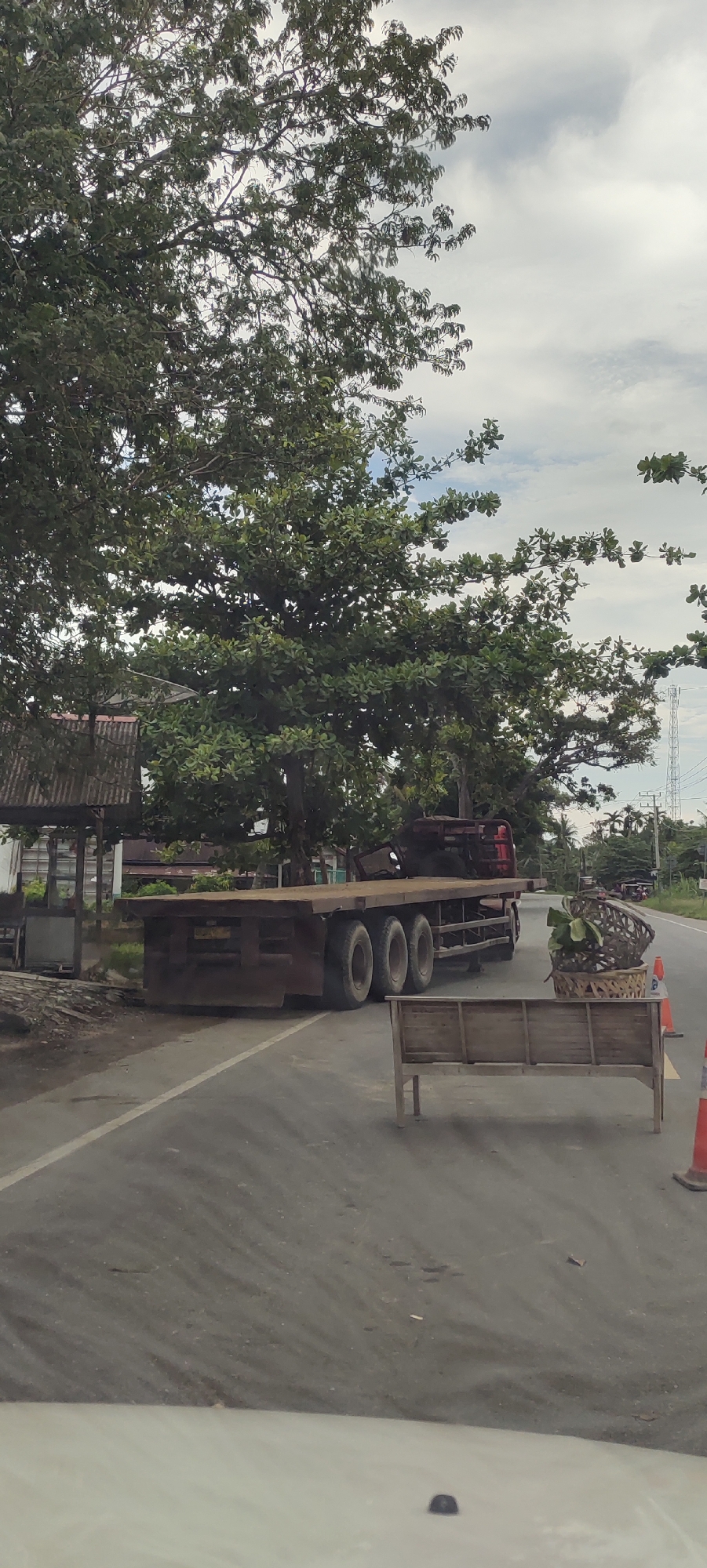 A large, flatbed truck with a red cab sits partially parked on the side of a paved road in Matangpanyang, Indonesia. The truck bed appears empty. To the right of the truck, in the road, a wooden bench and a basket with vegetation are situated, flanked by orange traffic cones. The scene is partly obscured by lush, green trees. A small structure with a corrugated metal roof is visible to the left of the truck. Power lines are visible in the background to the right. The sky is overcast. The photo appears to have been taken from inside a vehicle.