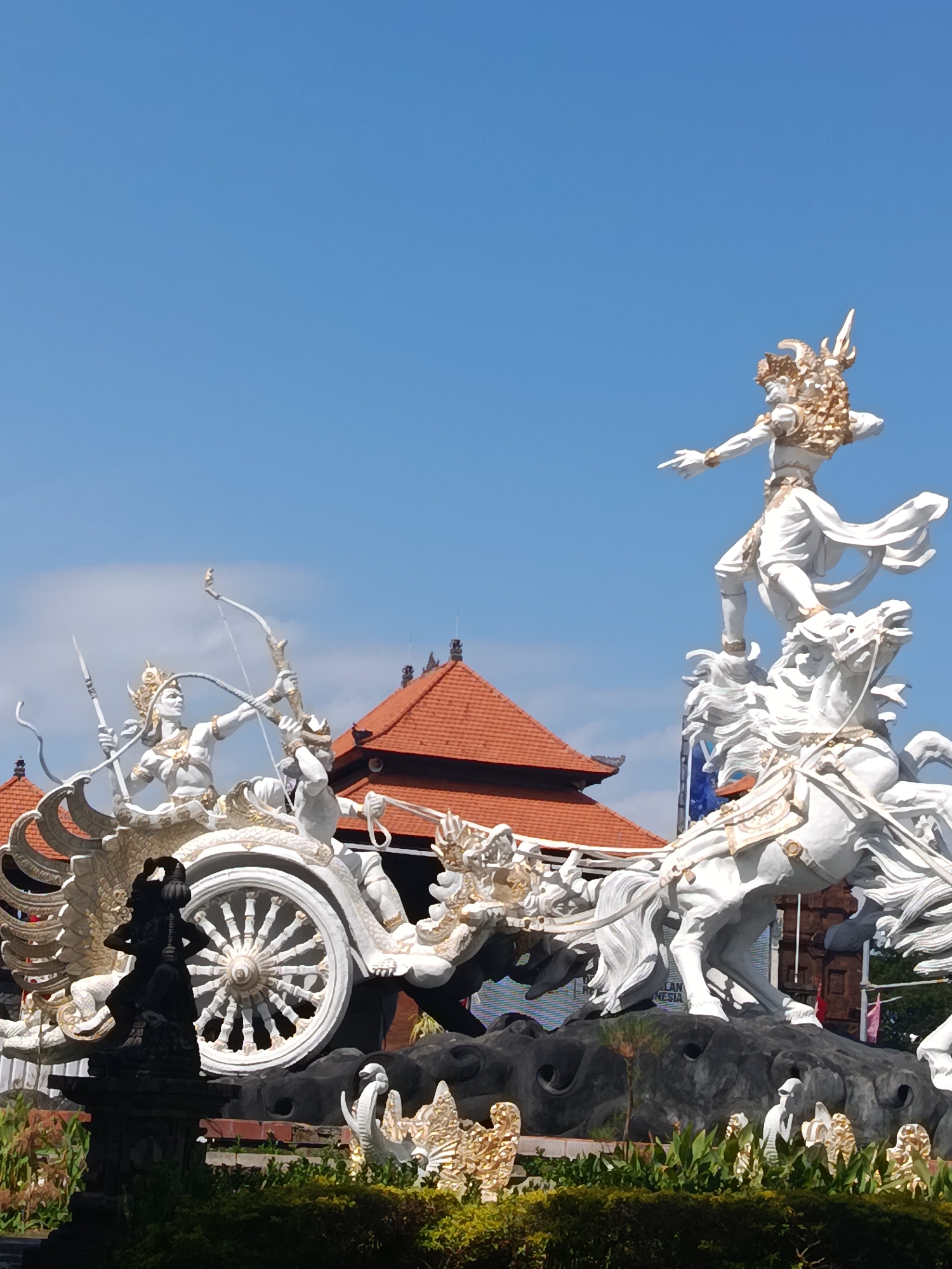 This is a medium shot of a monumental statue in Kuta, Indonesia. The statue depicts a chariot pulled by two horses, with figures of warriors and divine beings. The primary focus is on the white and gold statues, rendered in a dynamic and elaborate style.
In the foreground, a dark, small statue of a figure stands to the left, with its hands on its hips, on a stone pedestal. Behind it, lush green foliage creates a natural border.
The central element of the scene is a large, ornate chariot. One warrior, dressed in white and gold, is seated in the chariot, drawing a bow. Another warrior stands behind him, also appearing to be engaged in combat. The chariot itself is intricately designed, with a large spoked wheel as a prominent feature. The chariot is drawn by two white horses, which are depicted in a powerful, rearing stance.
Dominating the right side of the statue is another large white and gold figure, standing on the back of one of the horses, with one arm extended. This figure wears an elaborate headdress and flowing garments. The entire sculptural ensemble is rendered with fine detail, with gold accents highlighting the figures and their adornments.
In the background, a building with a distinctive red-tiled roof and traditional Indonesian architectural elements is visible. The sky is a clear, bright blue. The scene conveys a sense of epic action and grandeur, likely representing a mythological or historical event significant to Indonesian culture. The statue is situated in an outdoor setting, likely a public square or park, given its scale and prominent display.