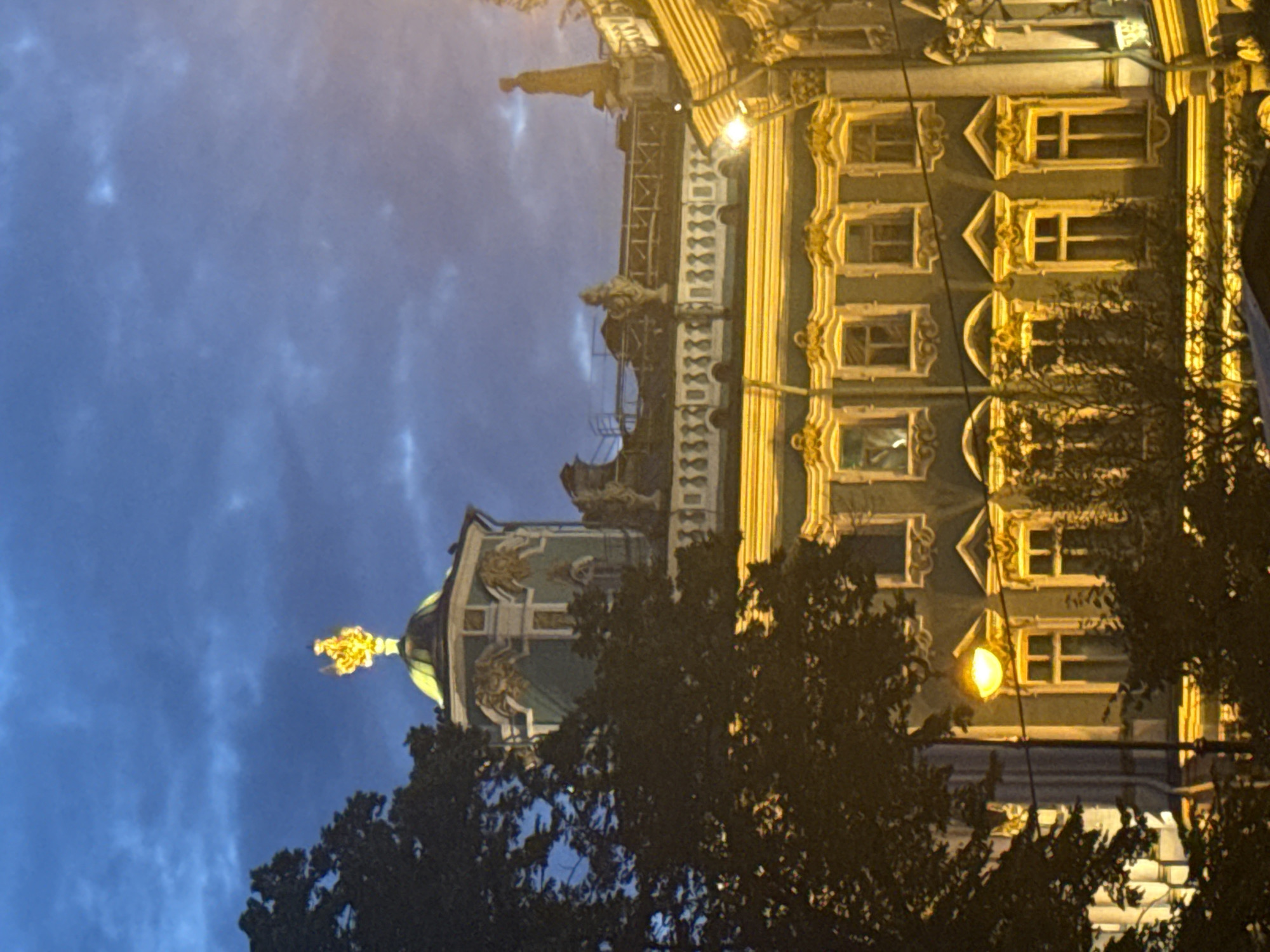 The image captures the facade of the Winter Palace in Saint Petersburg, Russia, at dusk. The sky is a deep blue with dramatic, dark clouds, suggesting twilight or early evening. The architecture is opulent and classical, with a prominent golden dome and intricate golden detailing on the upper levels. Several windows with ornate frames are visible, illuminated from within or by exterior lights, casting a warm glow. A large tree with dark foliage partially obscures the left side of the building. A streetlamp with a bright, yellow light is positioned in the foreground, adding to the overall illumination of the scene. The overall impression is one of grandeur and historical significance, bathed in the atmospheric light of twilight.