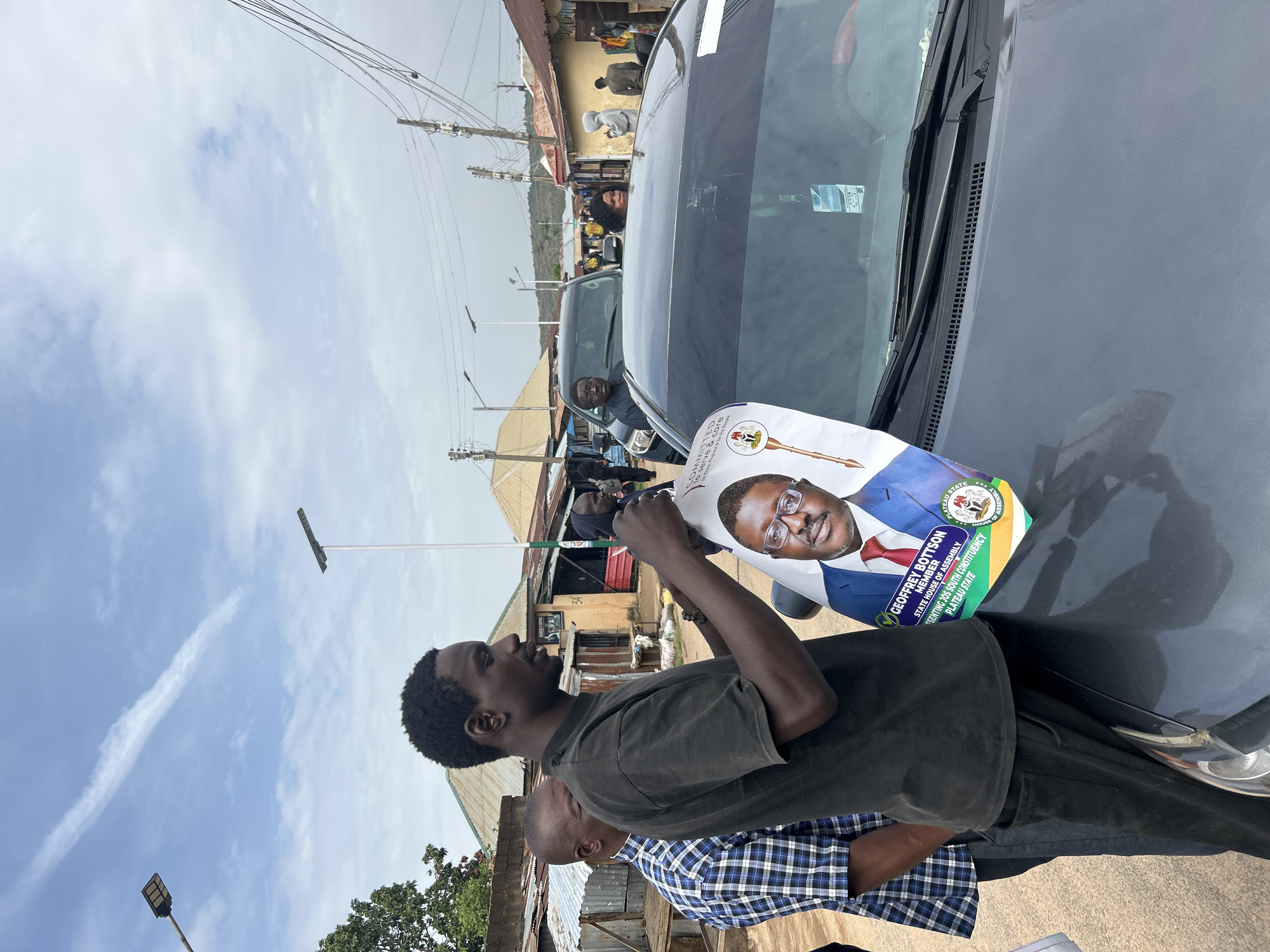 A man stands next to a black car in what appears to be an urban area. He is holding a large campaign flyer of Geoffrey Bottson, a candidate for the State House of Assembly representing Jos South Constituency in Plateau State. The flyer features Bottson's photo and the campaign slogan "Committed to serve & care for Peace, Progress & for the People." Another person in a blue and white plaid shirt is partially visible behind him. Street scenes with buildings and people are visible in the background. There are wires overhead.
