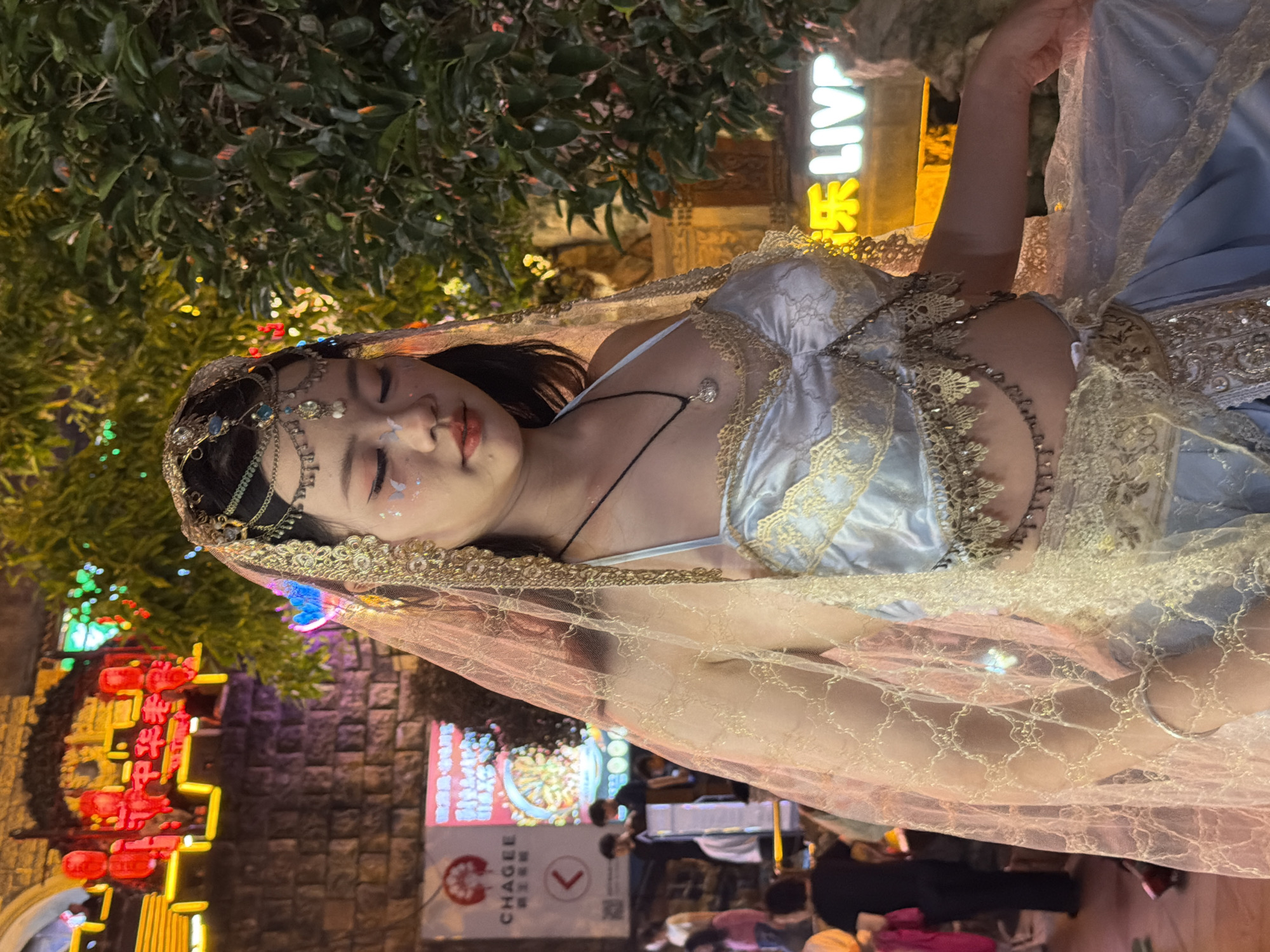 In a nighttime outdoor scene in the Yuzhong District of China, a young woman wearing an elaborate, light blue and gold costume with a sheer veil is positioned centrally. Her eyes are closed, and she has delicate butterfly-shaped makeup on her eyelids. She is adorned with a jeweled headpiece and intricate gold lace on her costume, which includes a cropped top and a flowing skirt. The background features a brick wall with neon signs and red lanterns, suggesting a commercial or entertainment area. Green foliage is visible to the right. In the background, several people are blurred, indicating they are not the main focus of the image. The overall atmosphere is festive and theatrical.